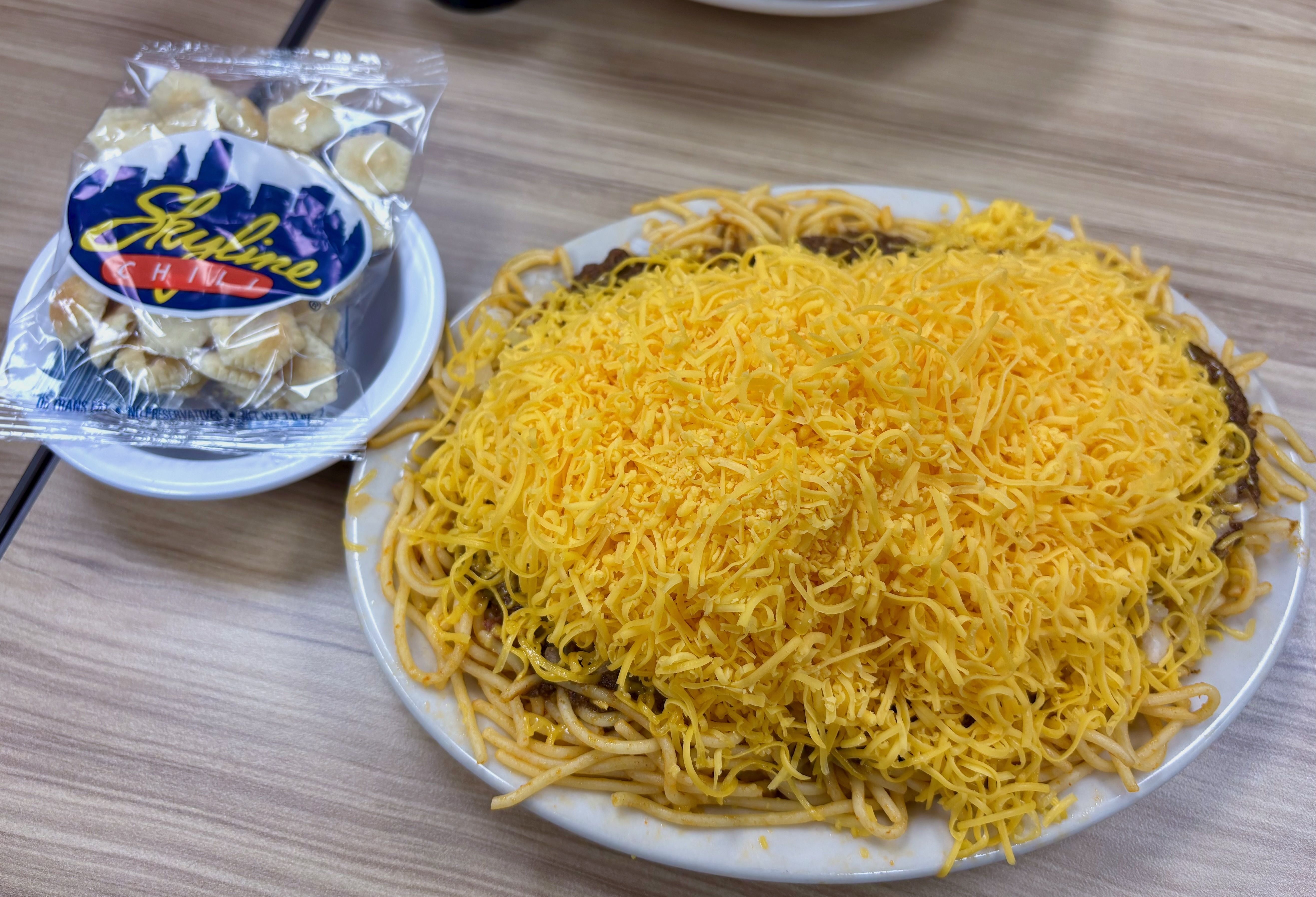 A small bowl with a bag of crackers that says Skyline Chili and to the right a big plate of spaghetti covered with chili and cheese.