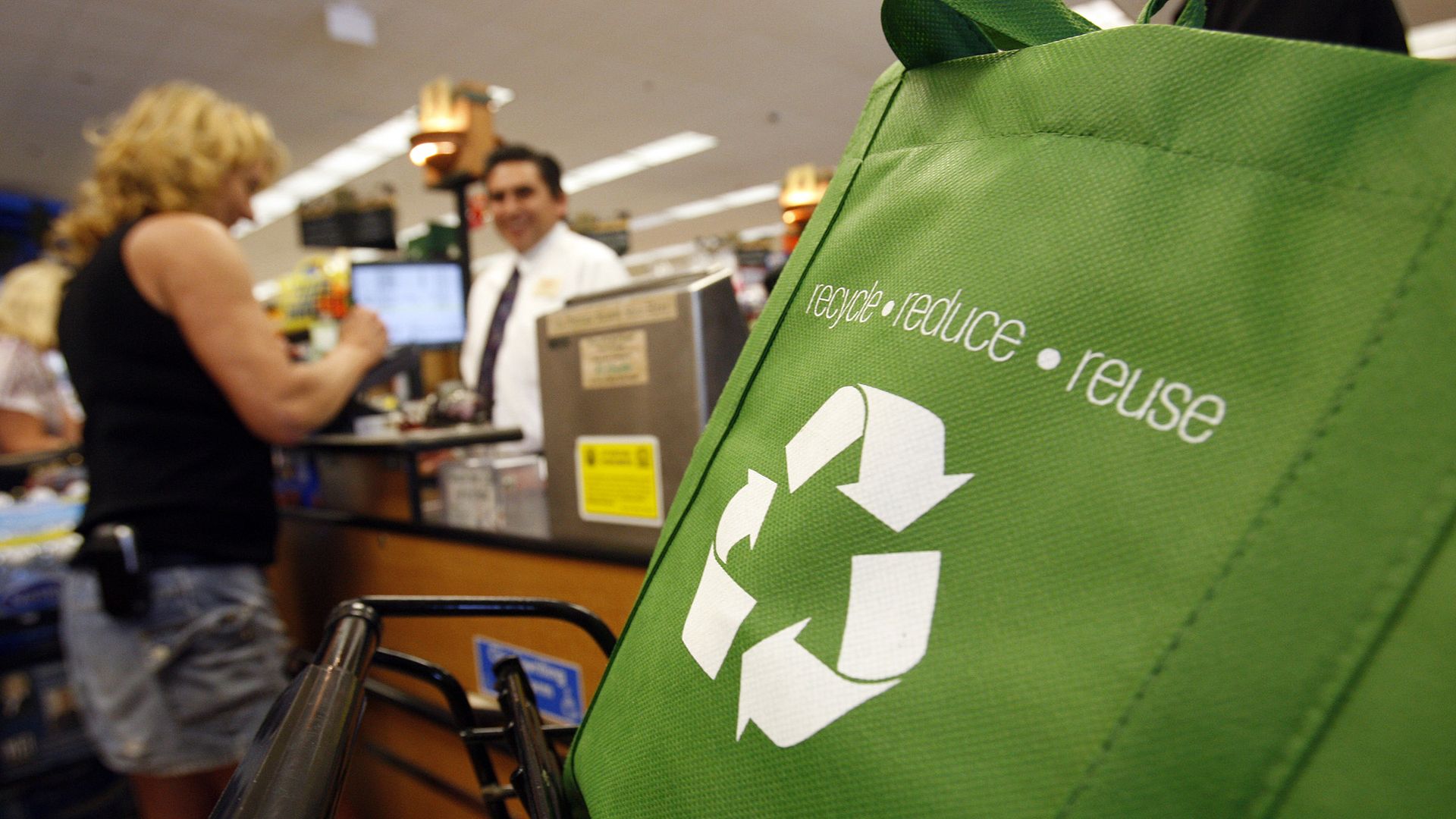 Woman checks out in grocery store in background, with reduce recycle reuse green bag in front.