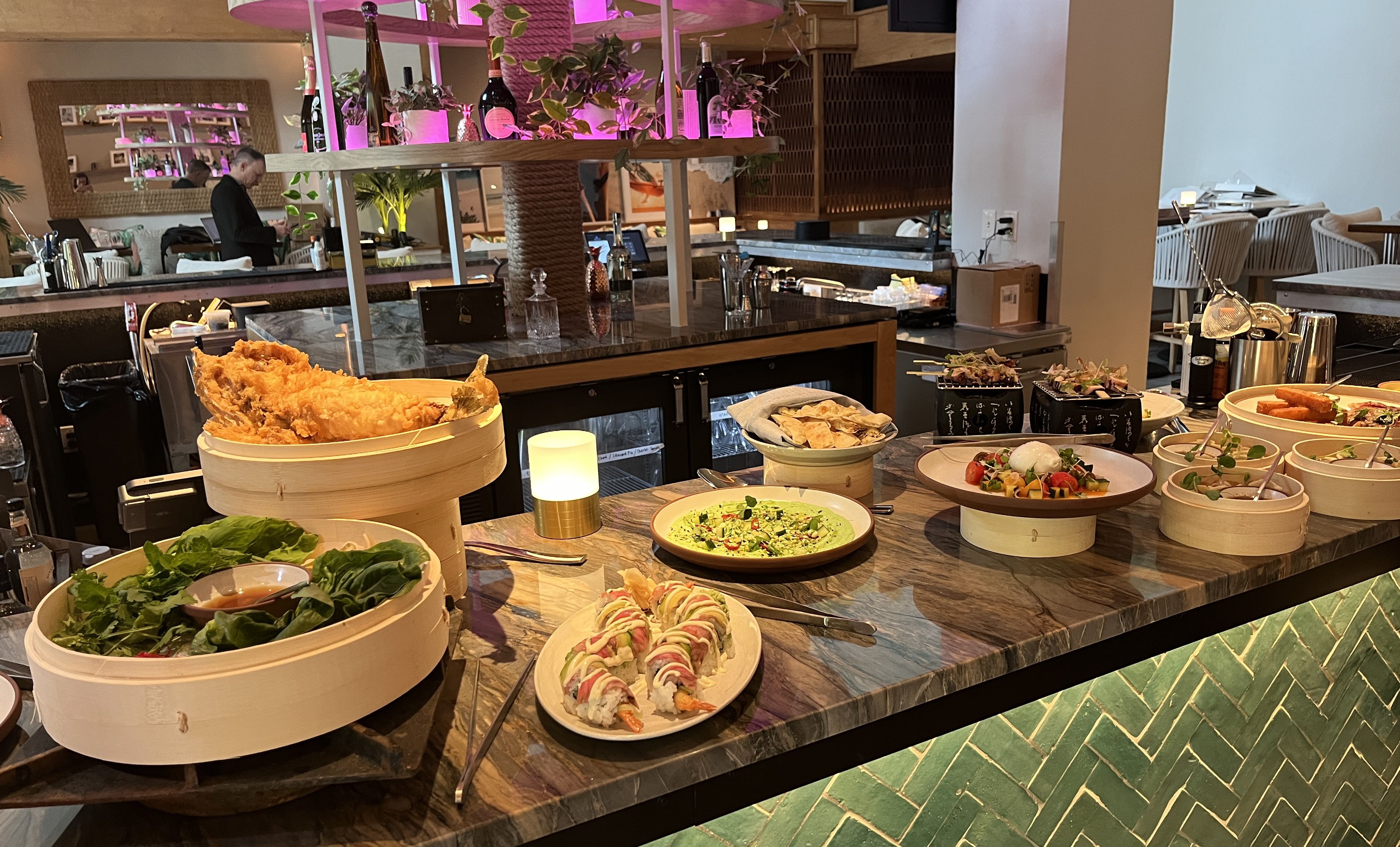Variety of colorful dishes including sushi, tempura, salads, and flatbreads arranged on a marble counter in a modern restaurant with plants and pink lighting in the background.