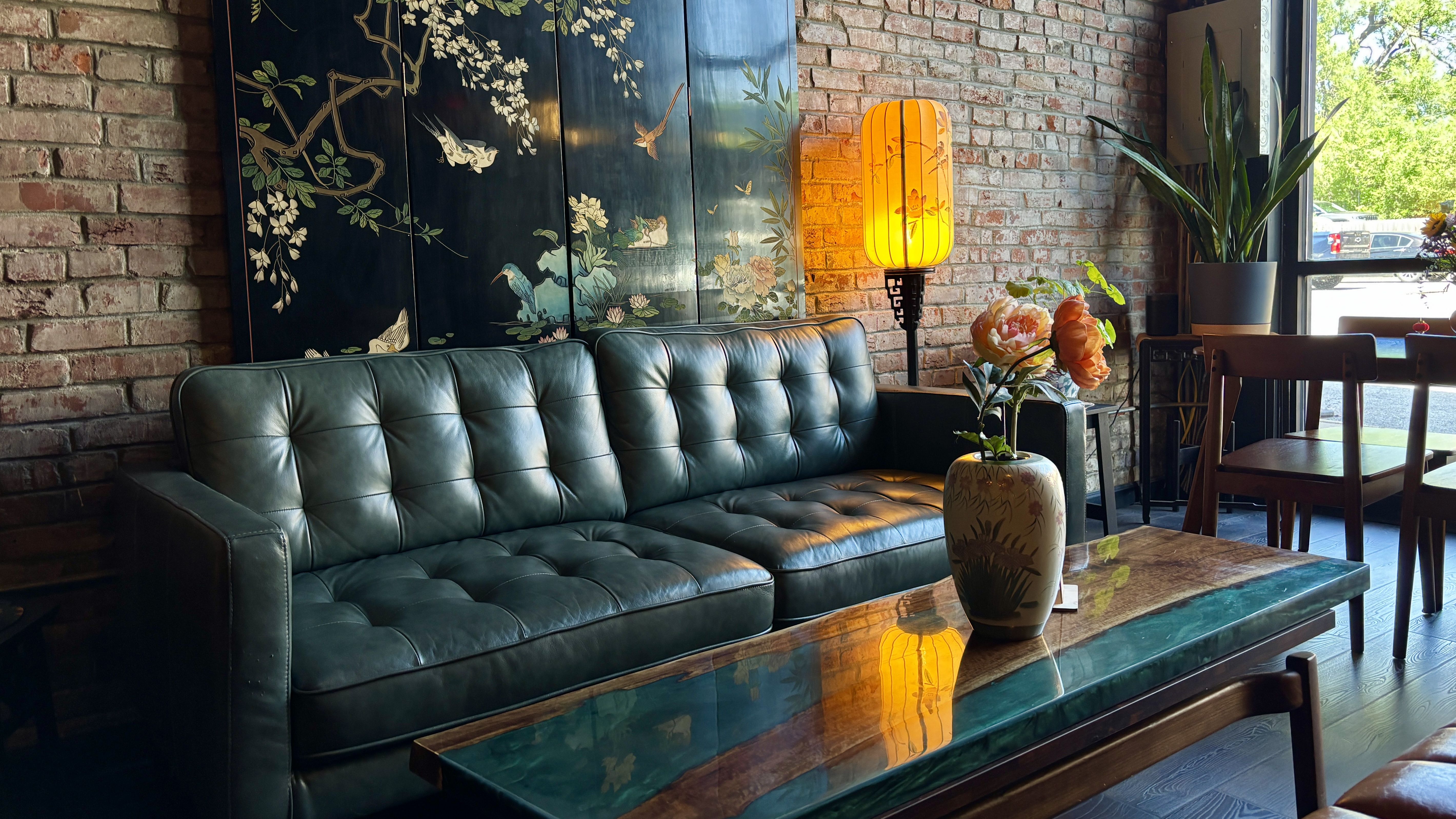 Cozy lounge with a teal tufted leather sofa, a blue decorative screen with birds and blossoms, a warm yellow lamp, potted plants, and brick walls; a window view of greenery outside.
