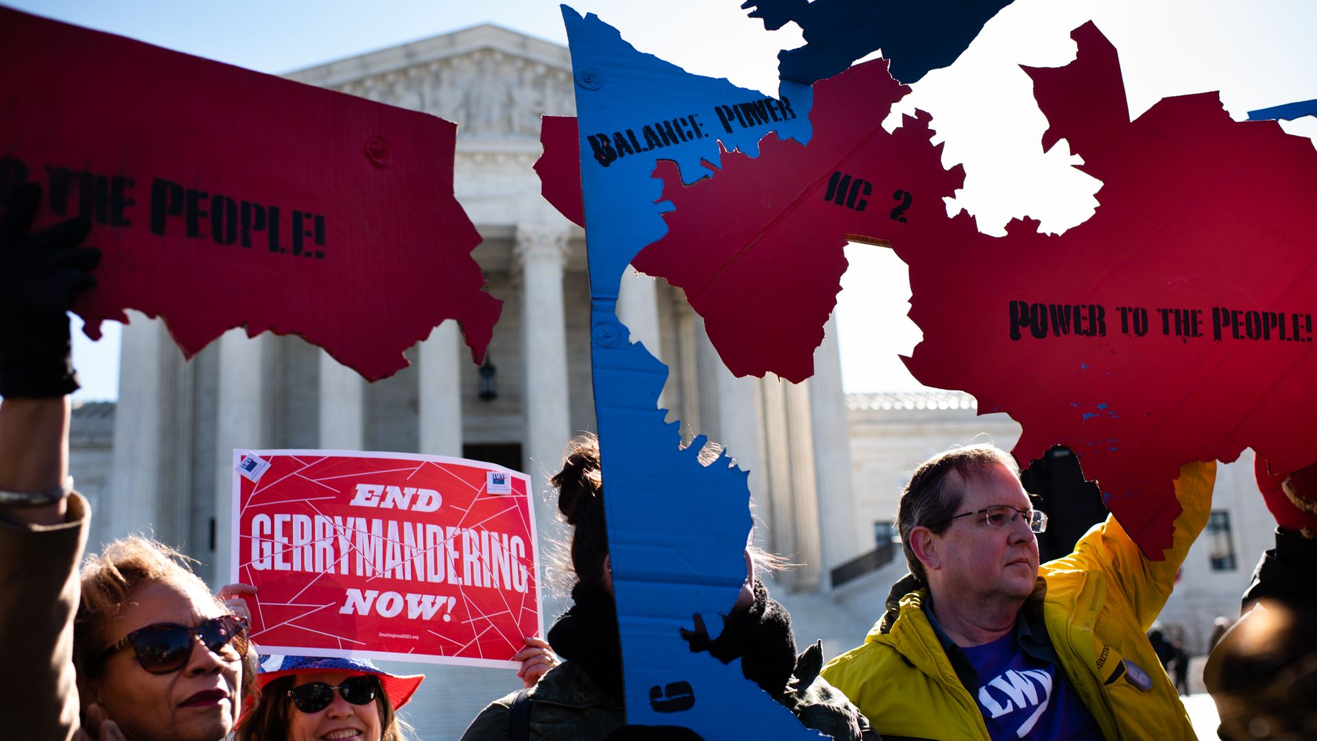 In this image, people in coats hold up signs that read "End Gerrymandering now" and "power to the people" while standing in front of the Supreme Court.