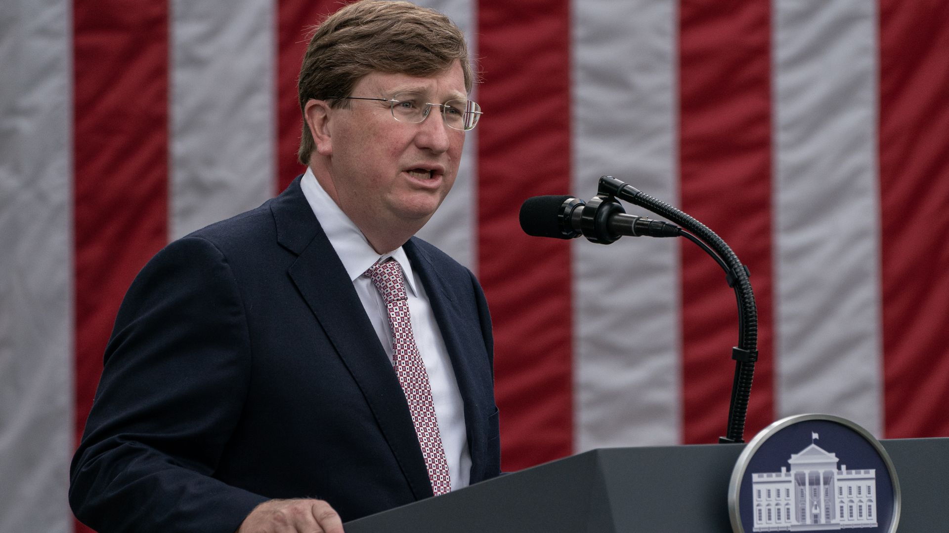 Photo of Tate Reeves speaking into a microphone from behind a podium with the American flag in the backdrop