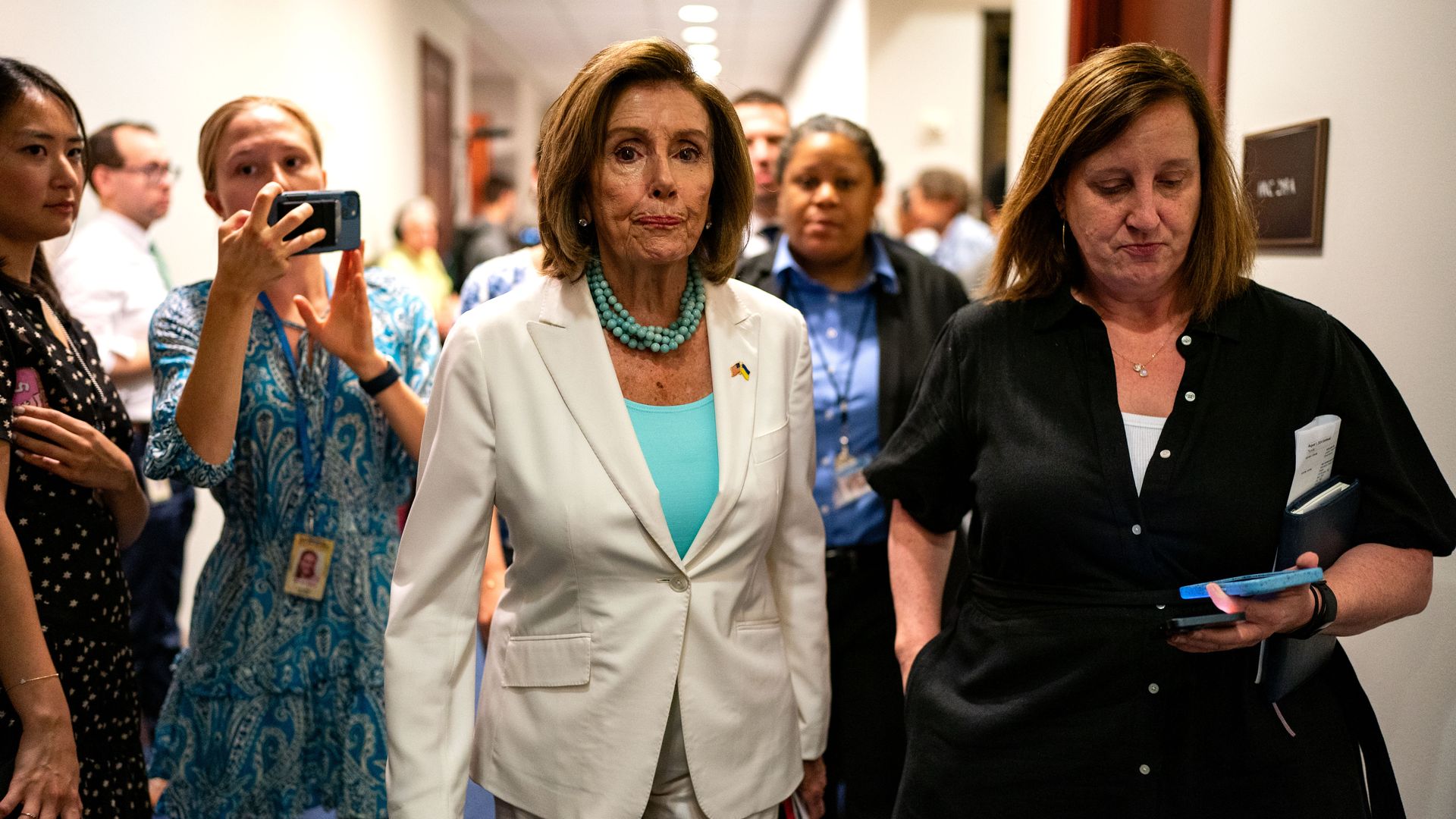 Former Speaker Nancy Pelosi, wearing a white suit, walking through a white hallway filled with reporters.
