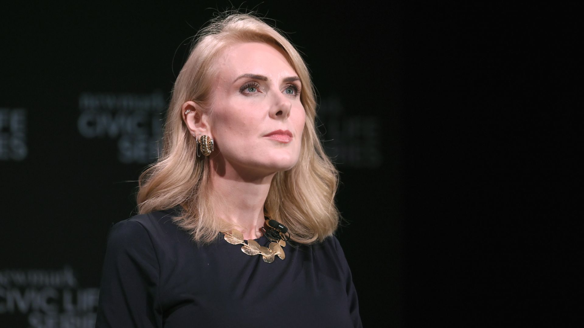 A blonde woman in a dark navy dress sits on a stage, wearing a gold leaf necklace and matching hoop earrings, with a microphone clipped to her neckline, gazing to the left against a dark backdrop.