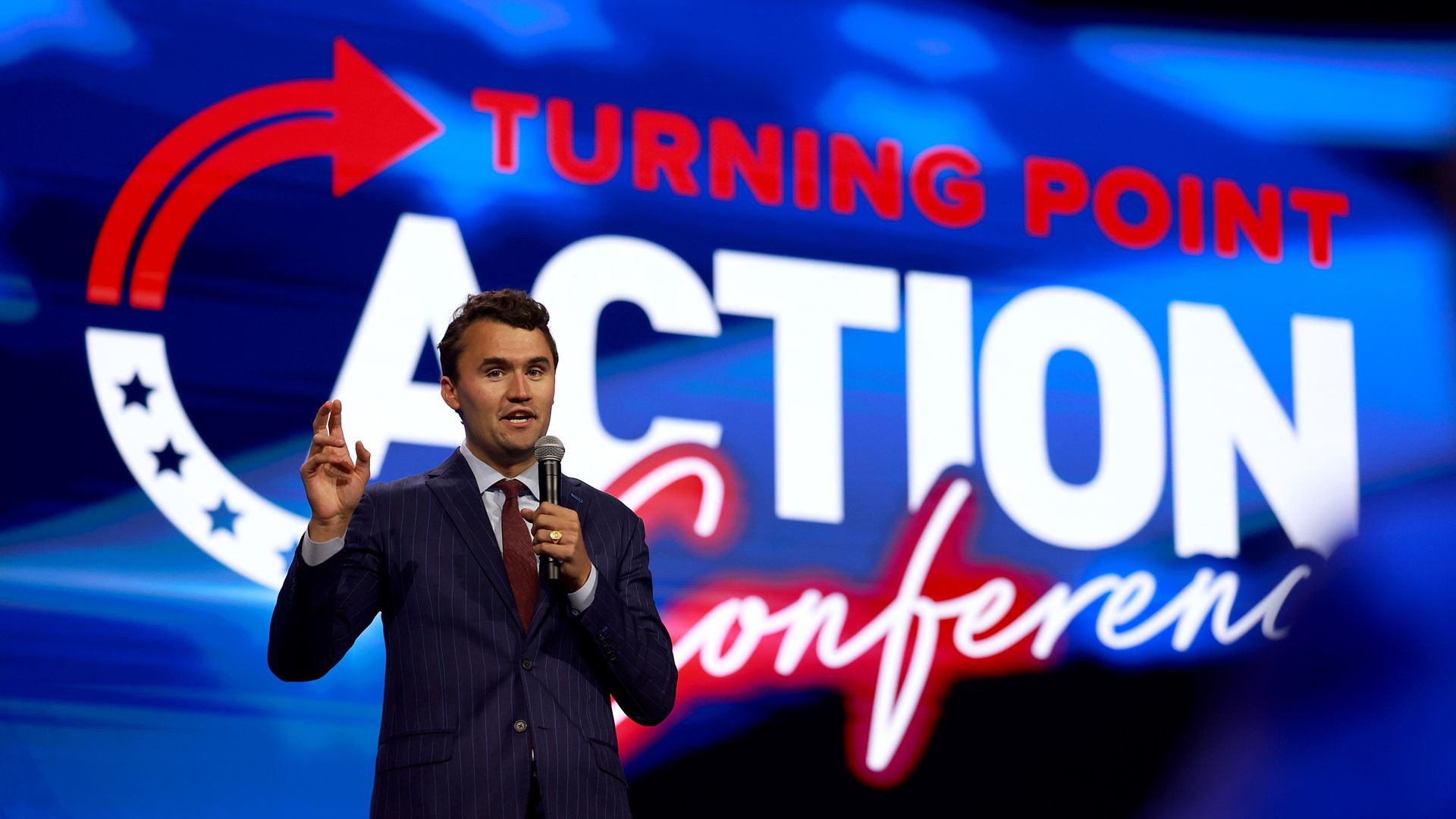 A man in a dark blue suit with a red tie and white shirt holds a microphone with his left hand and gestures with his right in front of a large digital display that reads "Turning Point Action Conference." 