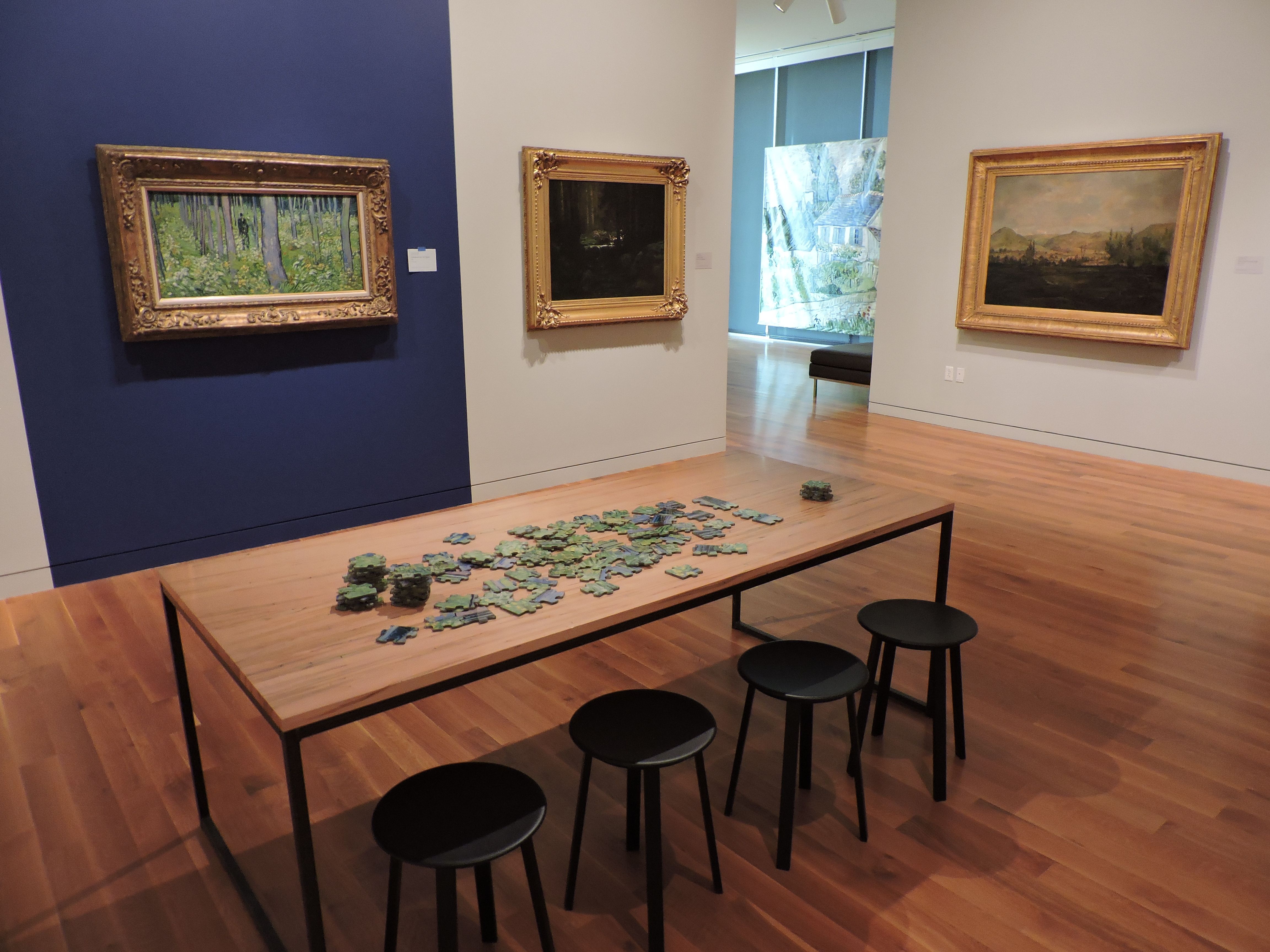A table with a large puzzle and stools sits in the middle of an art museum exhibit, with paintings on the walls