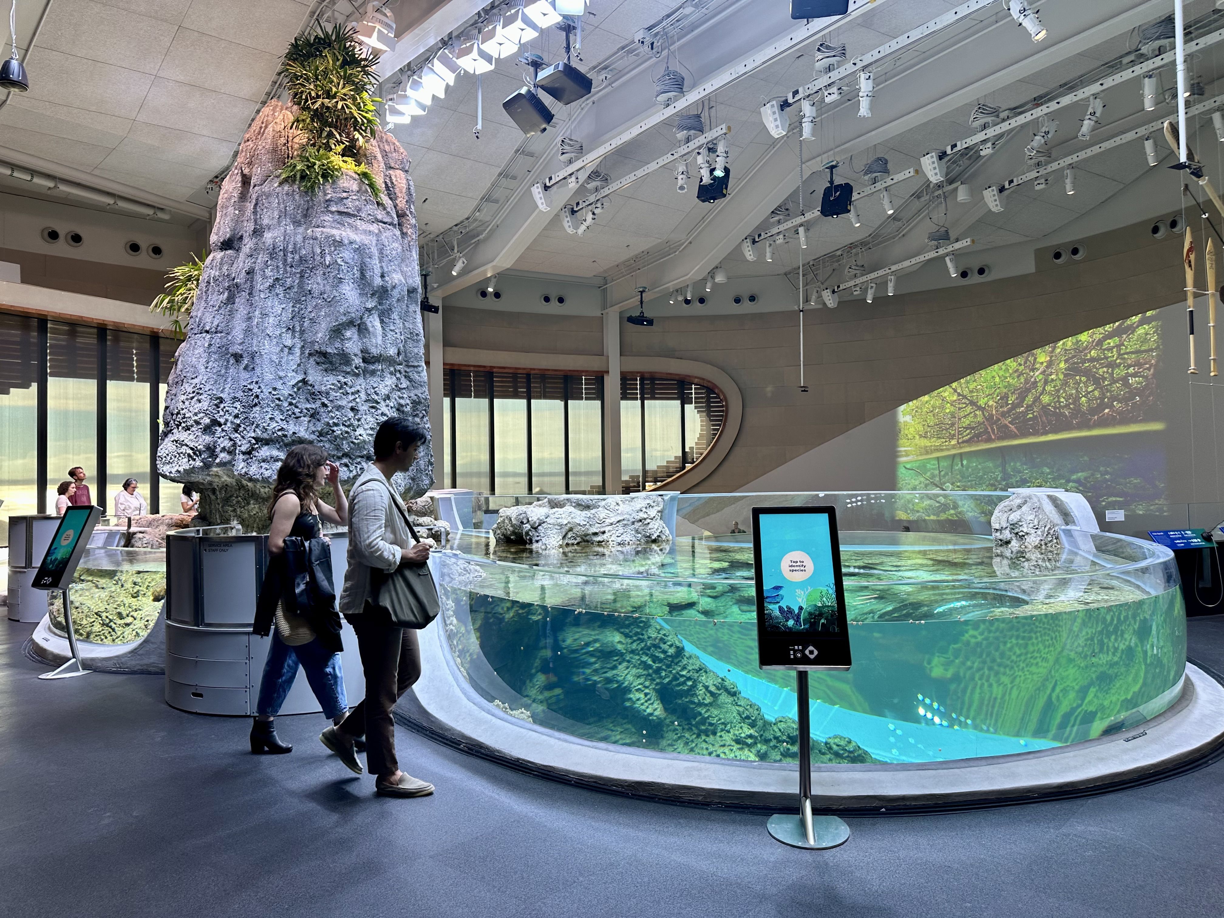 Two people walk past a rocky outcropping next to a pool of water encased in glass. An electronic screen sits at front to identify parts of the exhibit.