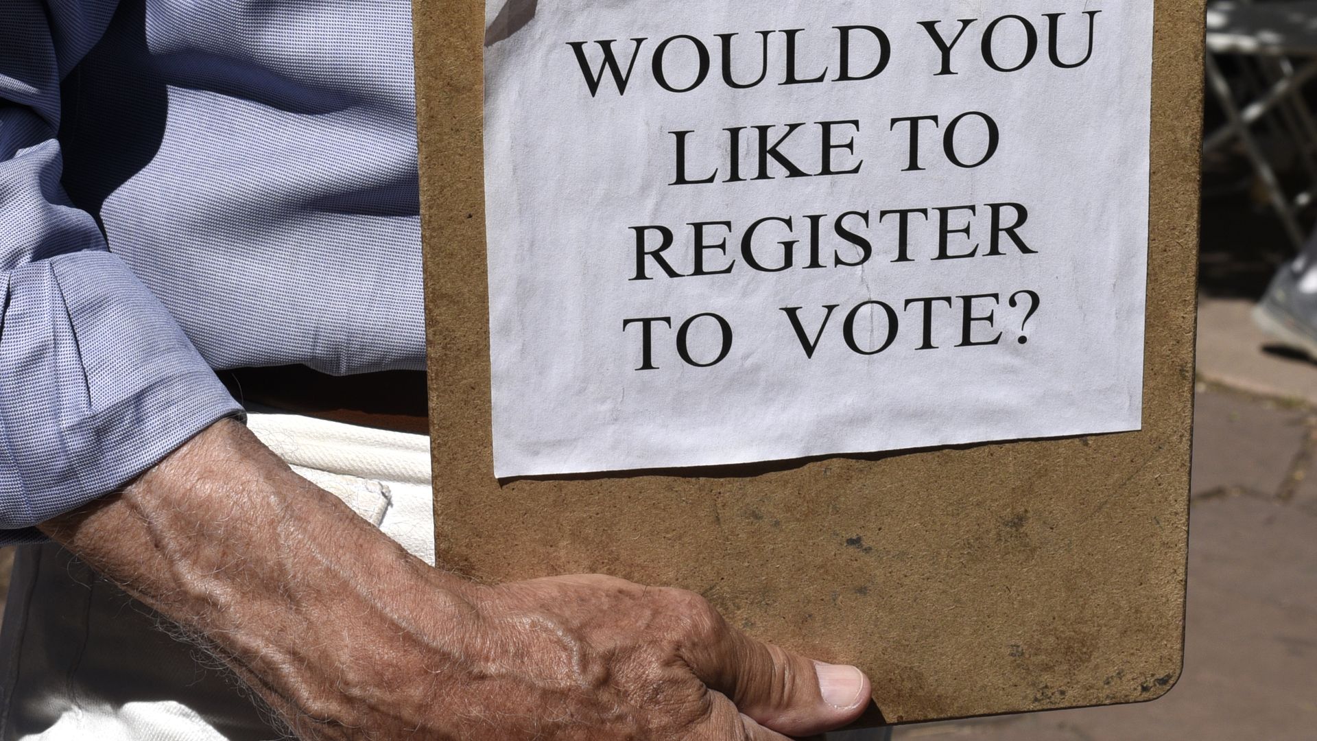 A voter registrar holding a clipboard that reads "Would you like to register to vote?"