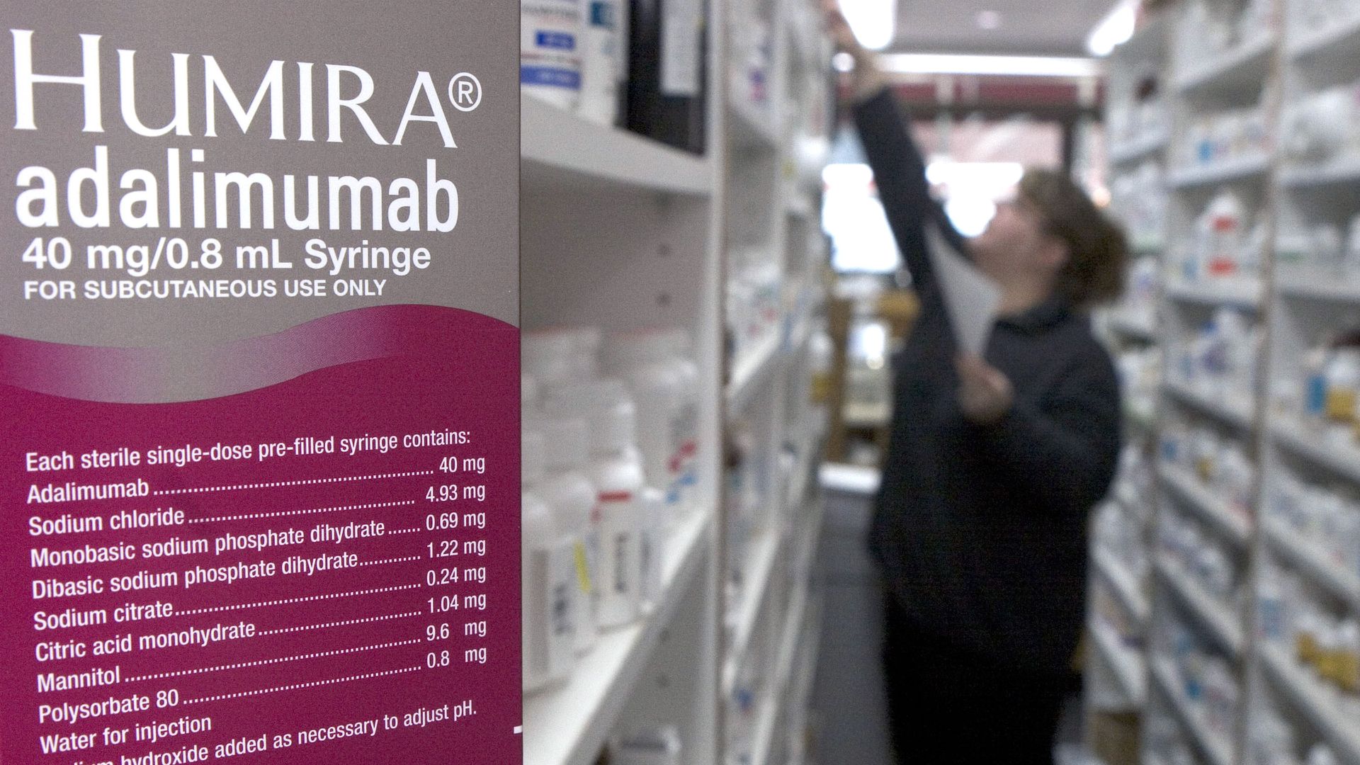 A box of the biosimilar Humira is shown in the foreground while a pharmacy staffer grabs a product of the shelf in the background.