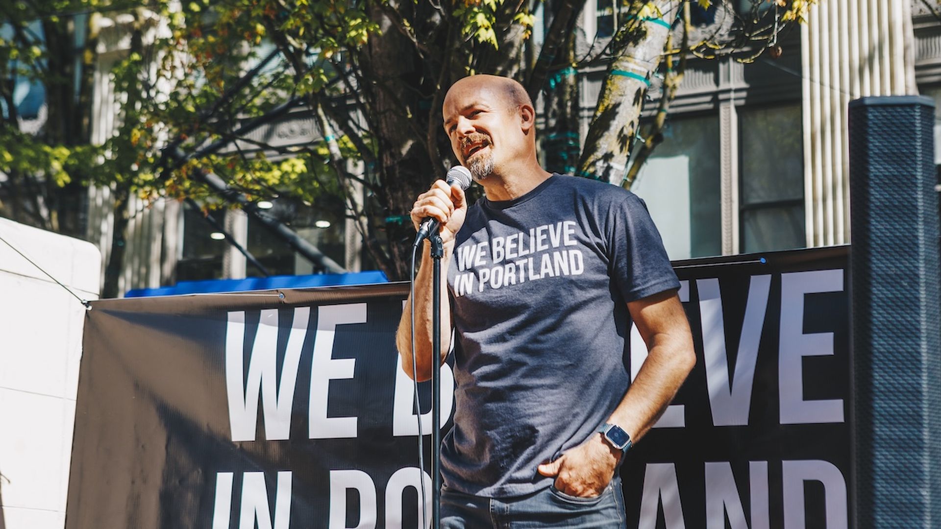 A photo of a person holding a microphone and wearing a shirt that reads "We believe in Portland"