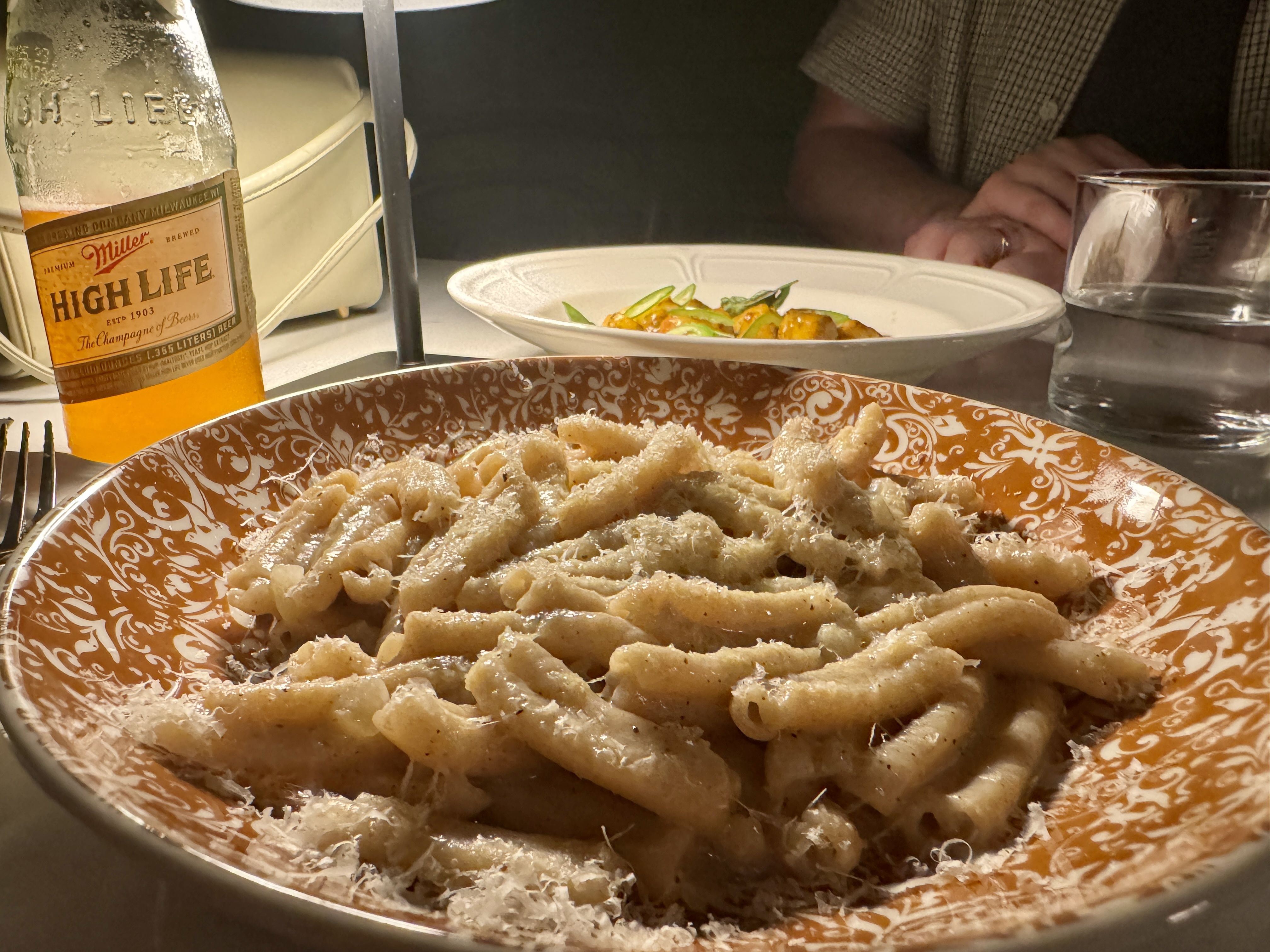 Plate of pasta with grated cheese on an ornate orange and white plate, a bottle of Miller High Life beer, a glass of water, and a person in a checkered shirt at a dining table.