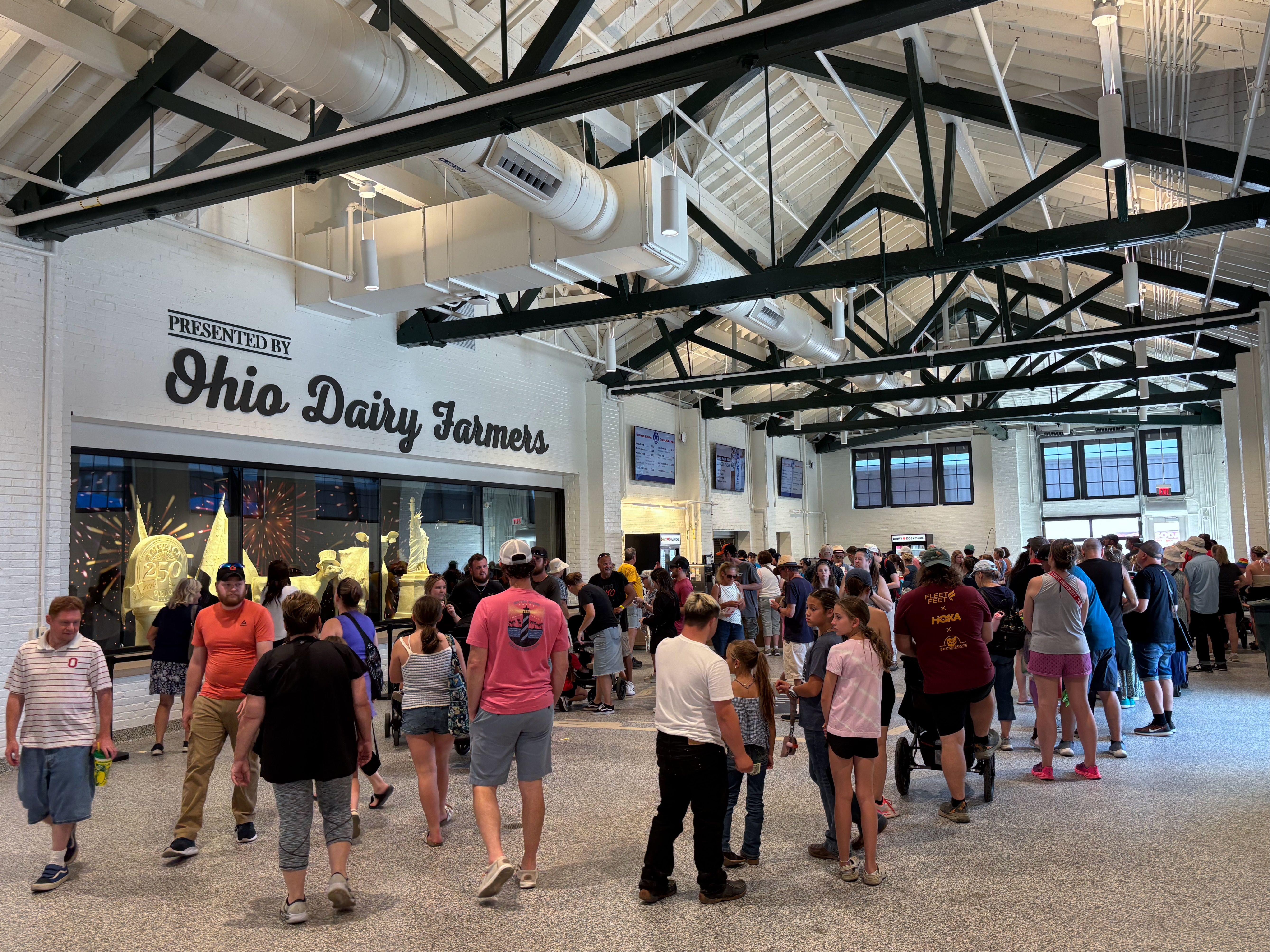 Inside a bustling hall with a large crowd, white brick walls, black steel trusses, and white ceiling ducts; a sign reads Presented by Ohio Dairy Farmers.