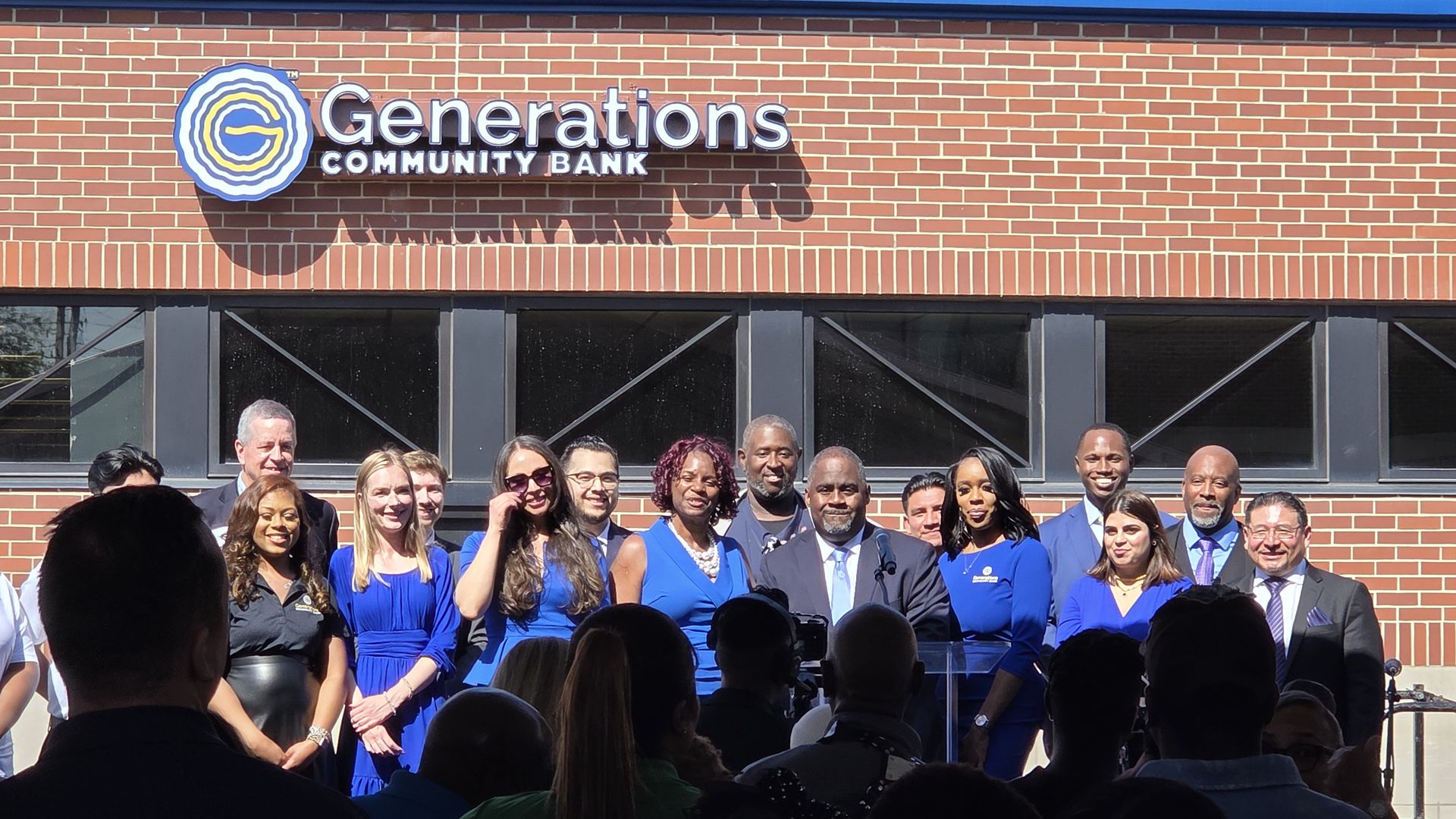 A diverse group of people in blue outfits pose in front of a brick building with a large sign that reads "Generations Community Bank" and a circular blue-yellow logo.
