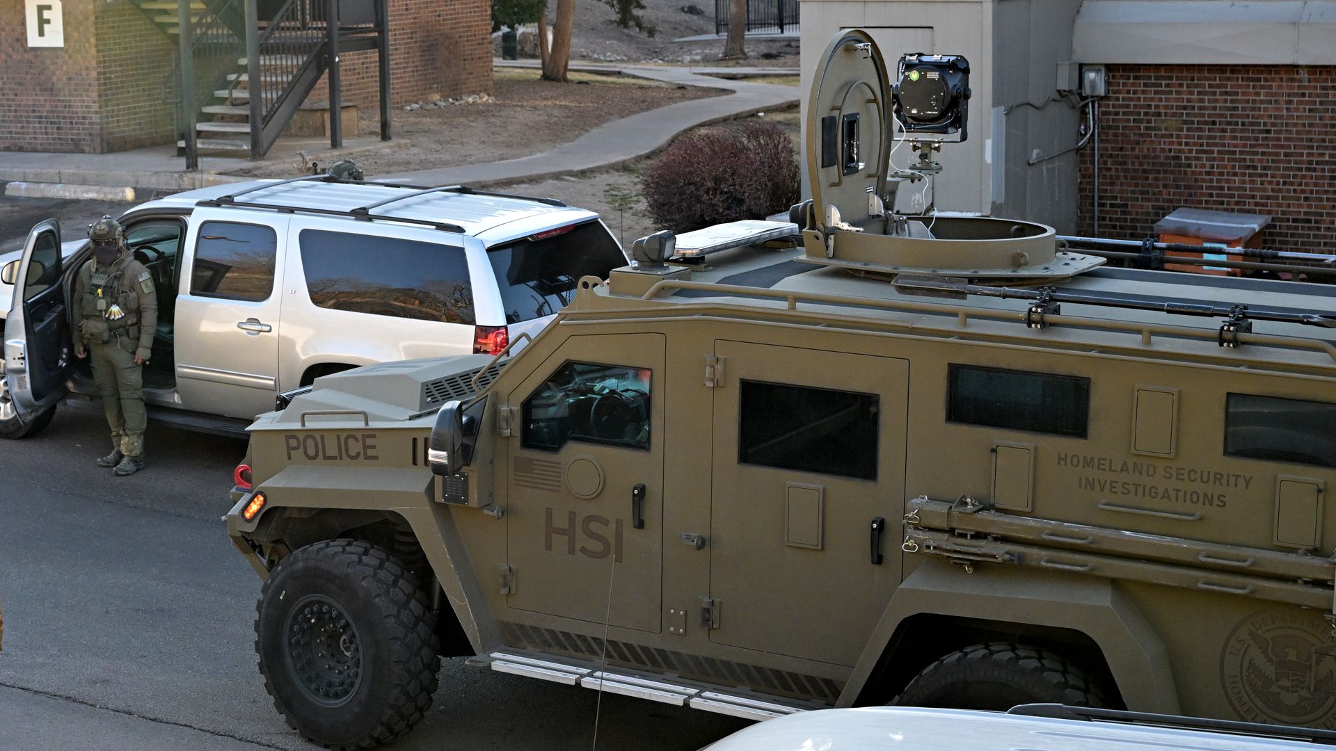 Child in a brown and blue jacket walking near a tan Homeland Security Investigations armored vehicle and a white SUV with an armed officer in tactical gear standing nearby in a parking lot.