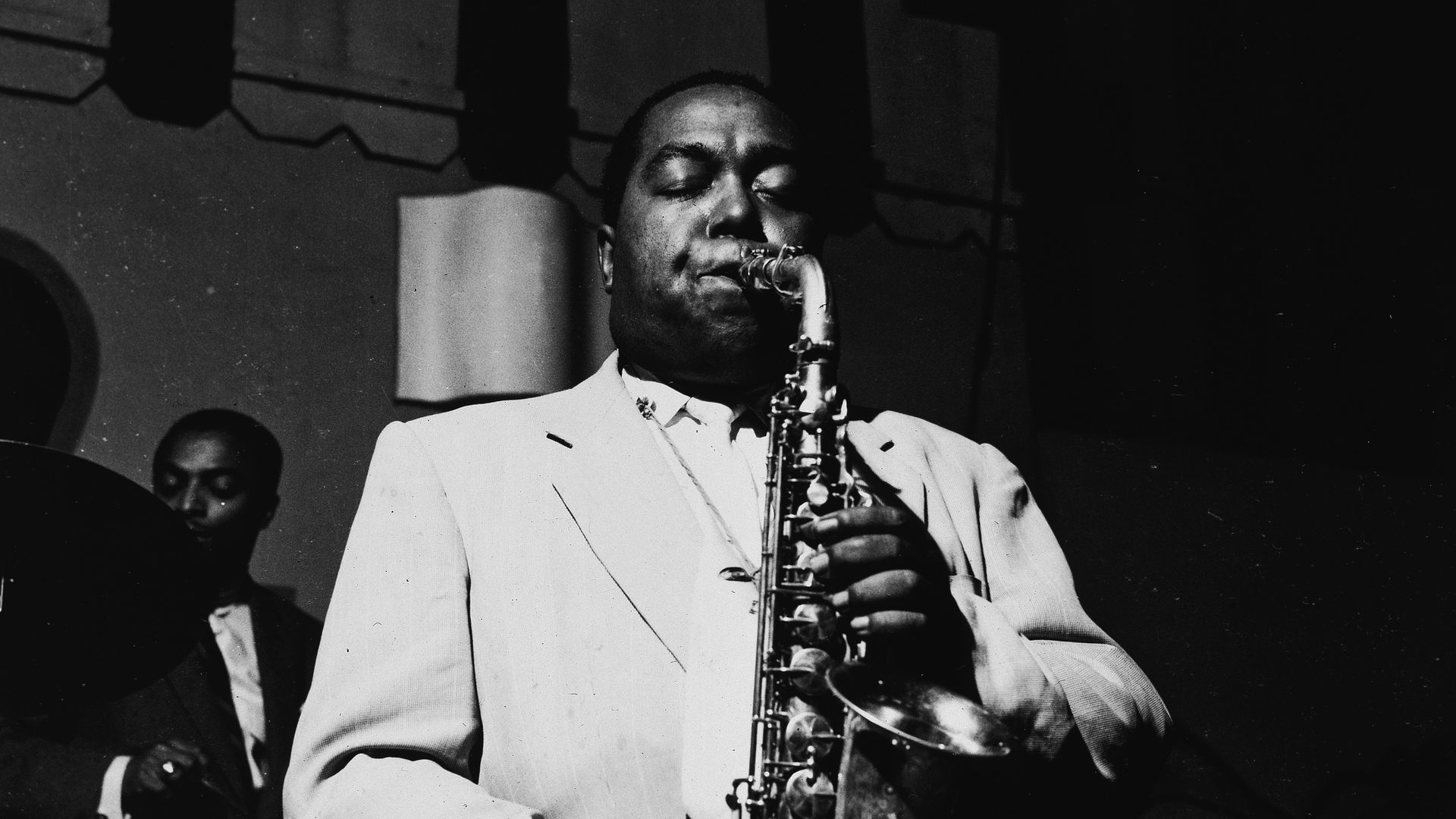 Black-and-white photo of a male jazz saxophonist in a light suit, eyes closed, playing on stage, with a drummer visible in the background.