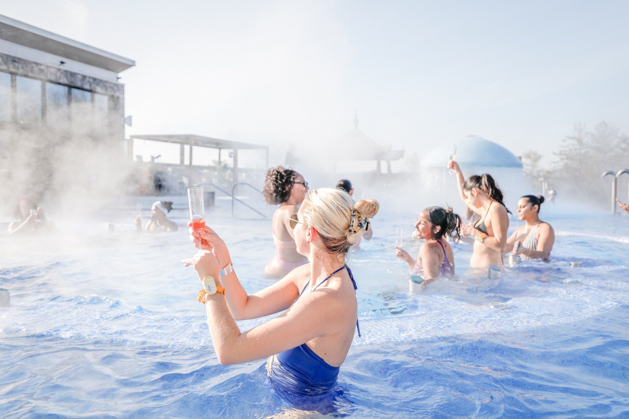 Group of women in swimsuits enjoying a steamy outdoor pool, holding drinks and socializing under bright daylight with a modern building and pavilion visible in the background.