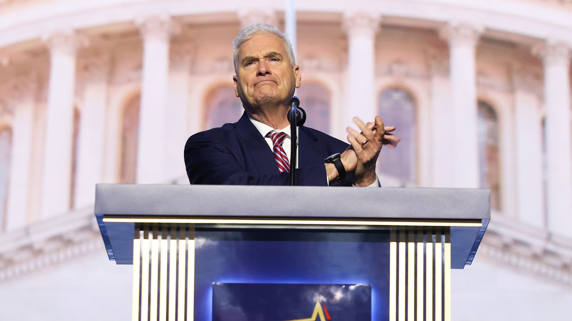 An older man in a dark suit and red striped tie speaks at a podium with a microphone; hands clasped. Behind him, tall columns and a Dallas Host Committee 2026 banner.