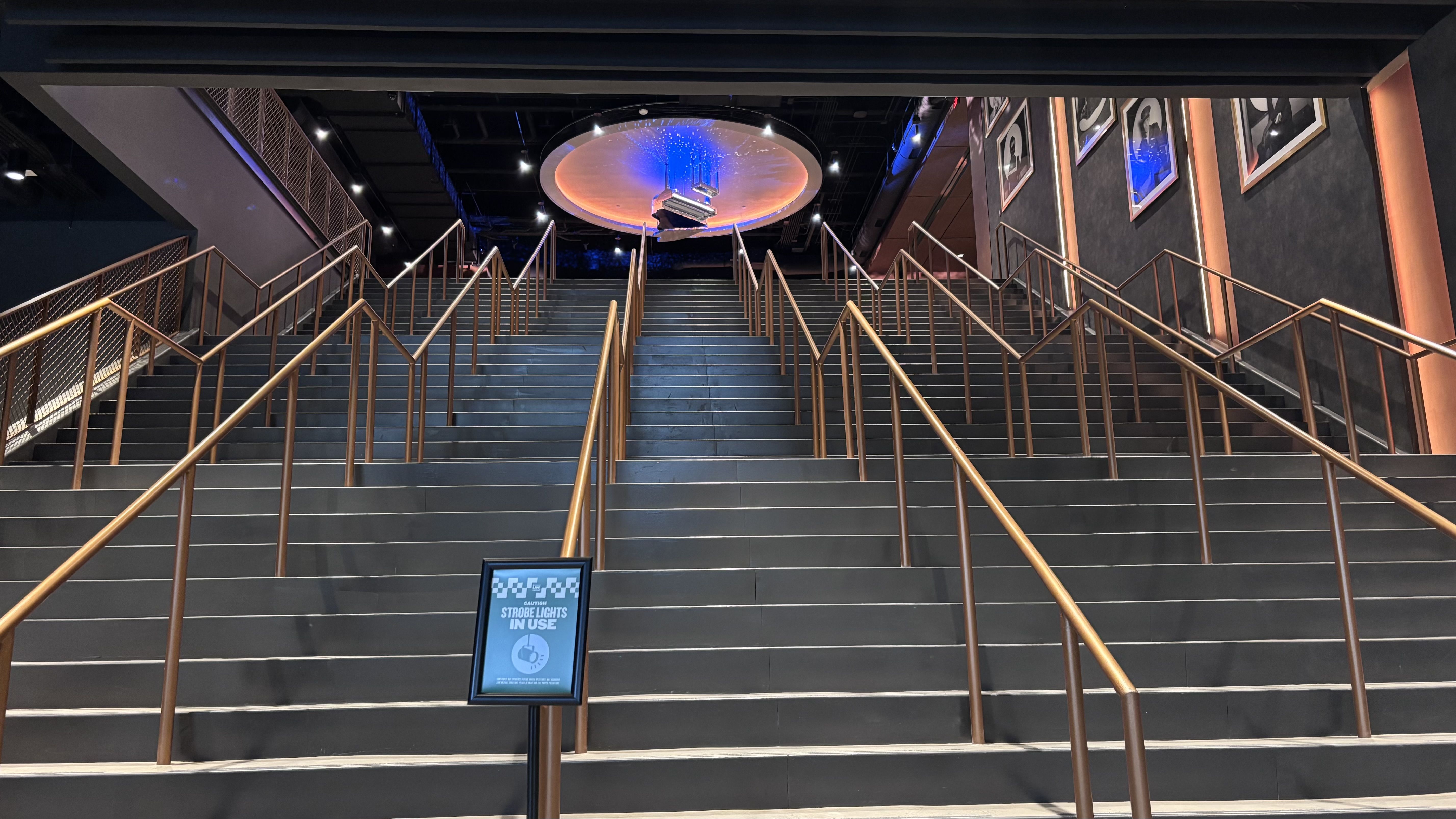 Wide indoor stairwell with bronze handrails, dark steps, and framed portraits on the right. A circular illuminated ceiling feature hangs above; a sign in the foreground reads "Strobe lights in use".