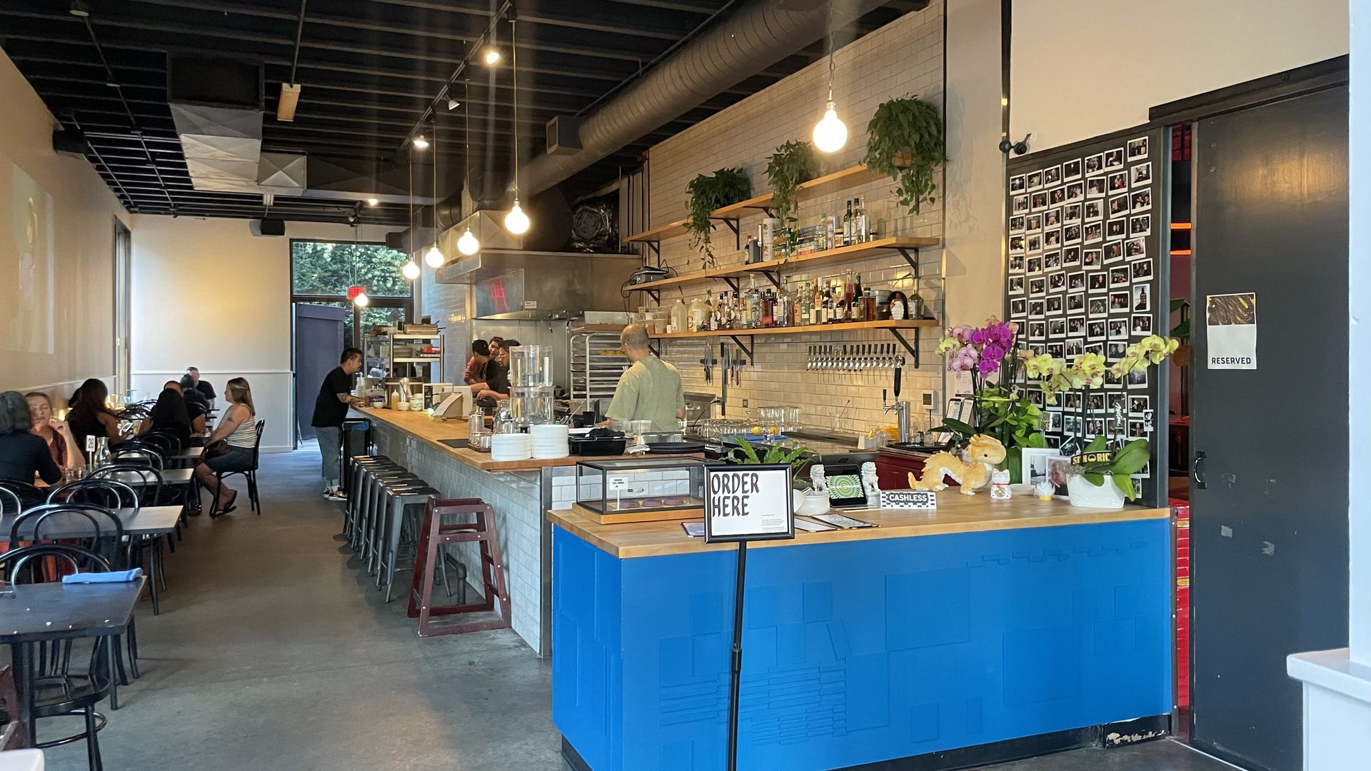 Interior of a modern cafe with a blue order counter, hanging light bulbs, shelves with bottles and plants, a black door with a "Reserved" sign, and customers seated at tables.