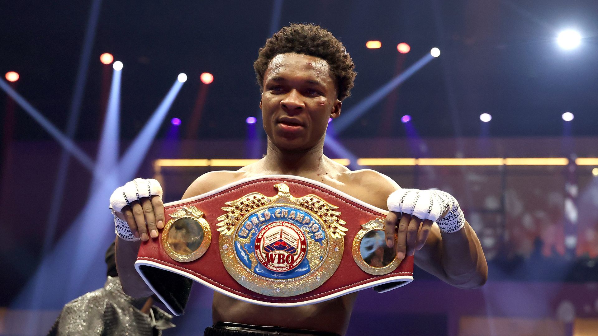 Boxer on stage, shirtless and sweaty, proudly displaying a red and gold WBO championship belt across his chest. Bright spotlights, wrapped fists, and a blurred crowd in the background.