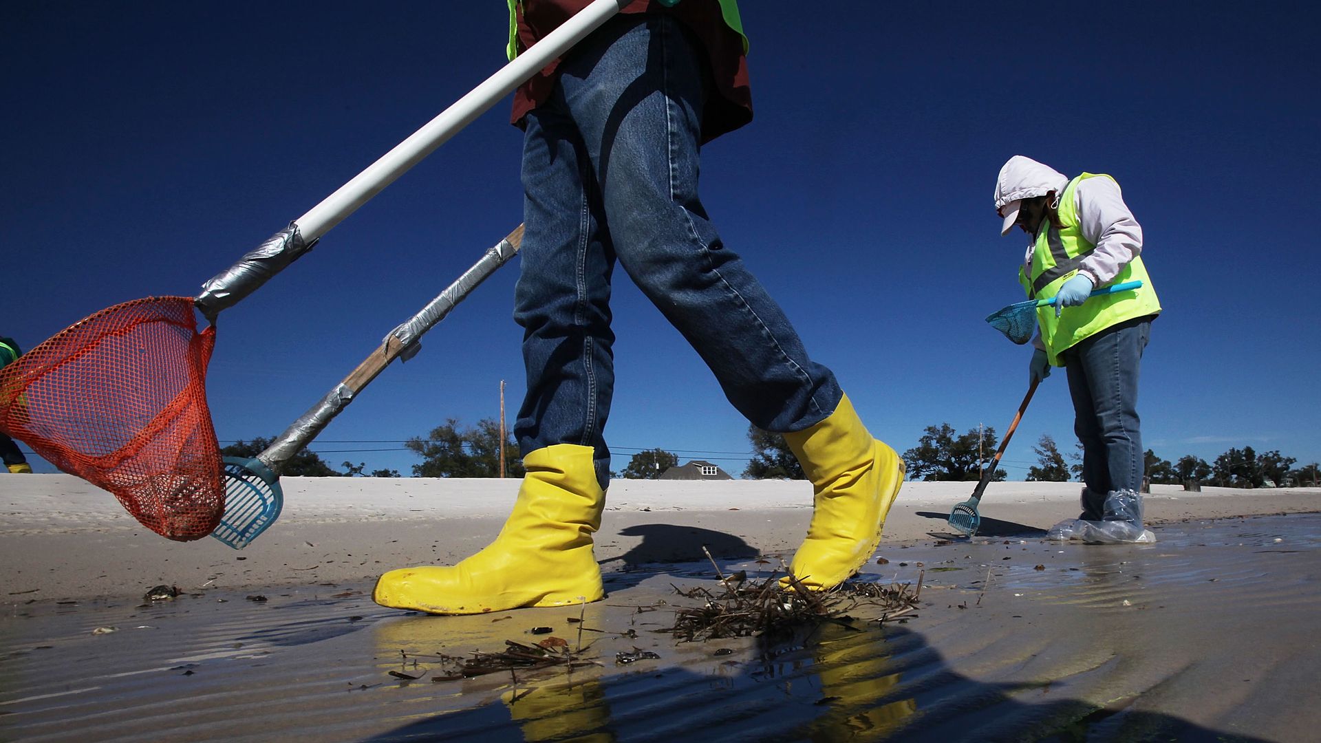 In this image, two men walk on a beach and clean tarballs.