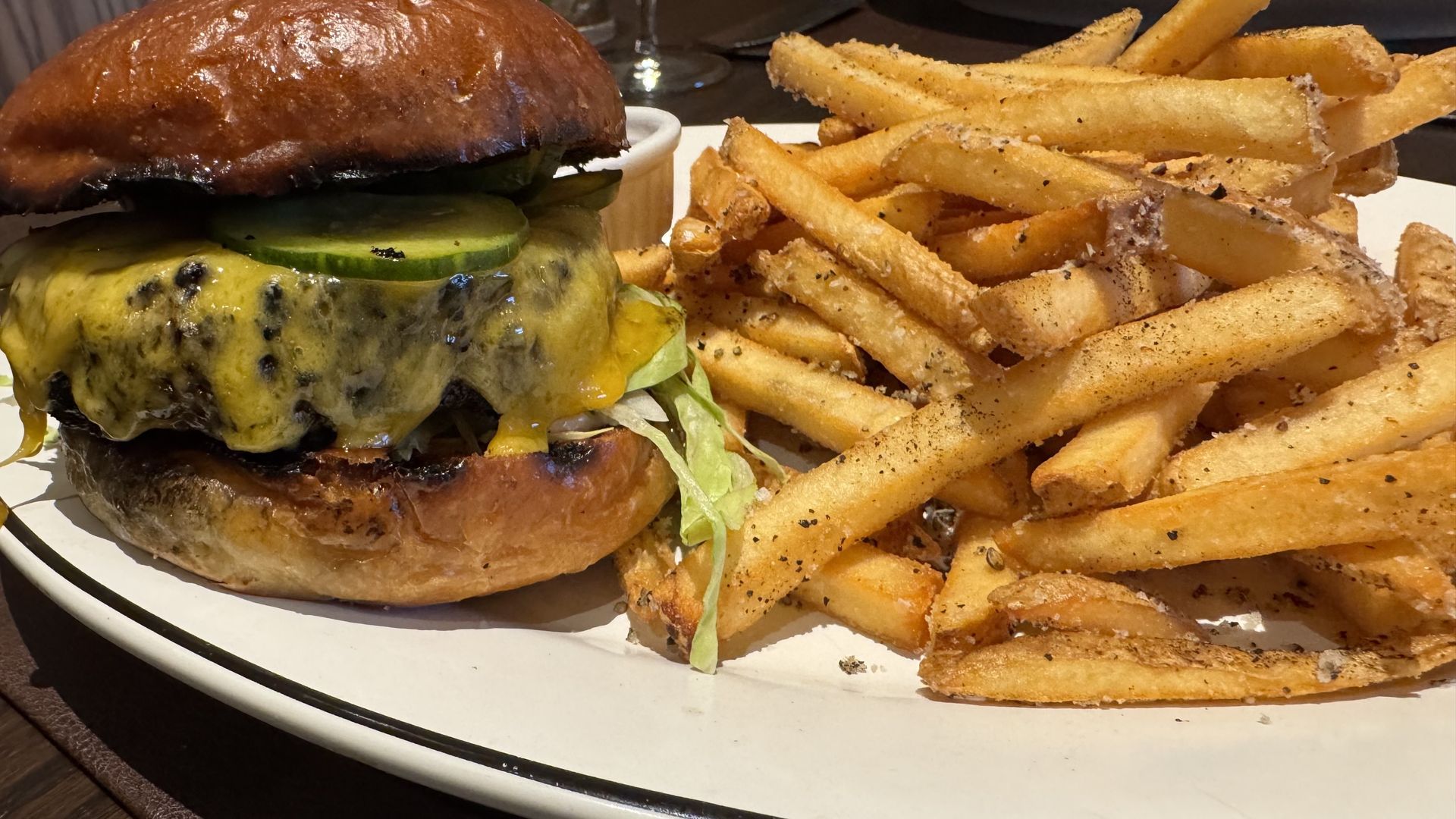 Juicy cheeseburger with melted cheddar, pickles, and lettuce in a glossy brioche bun, beside a heaping pile of seasoned french fries on a white plate.