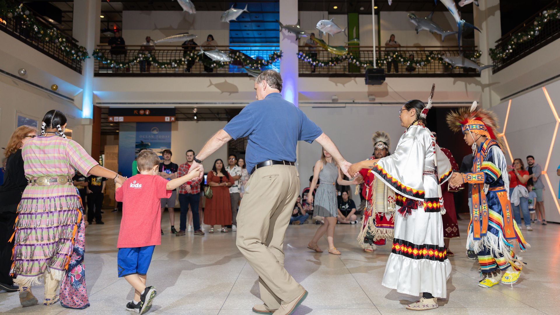 A diverse group of people holding hands dancing in a circle in a large indoor space with hanging fish models and an audience watching. Some wear Native American regalia.