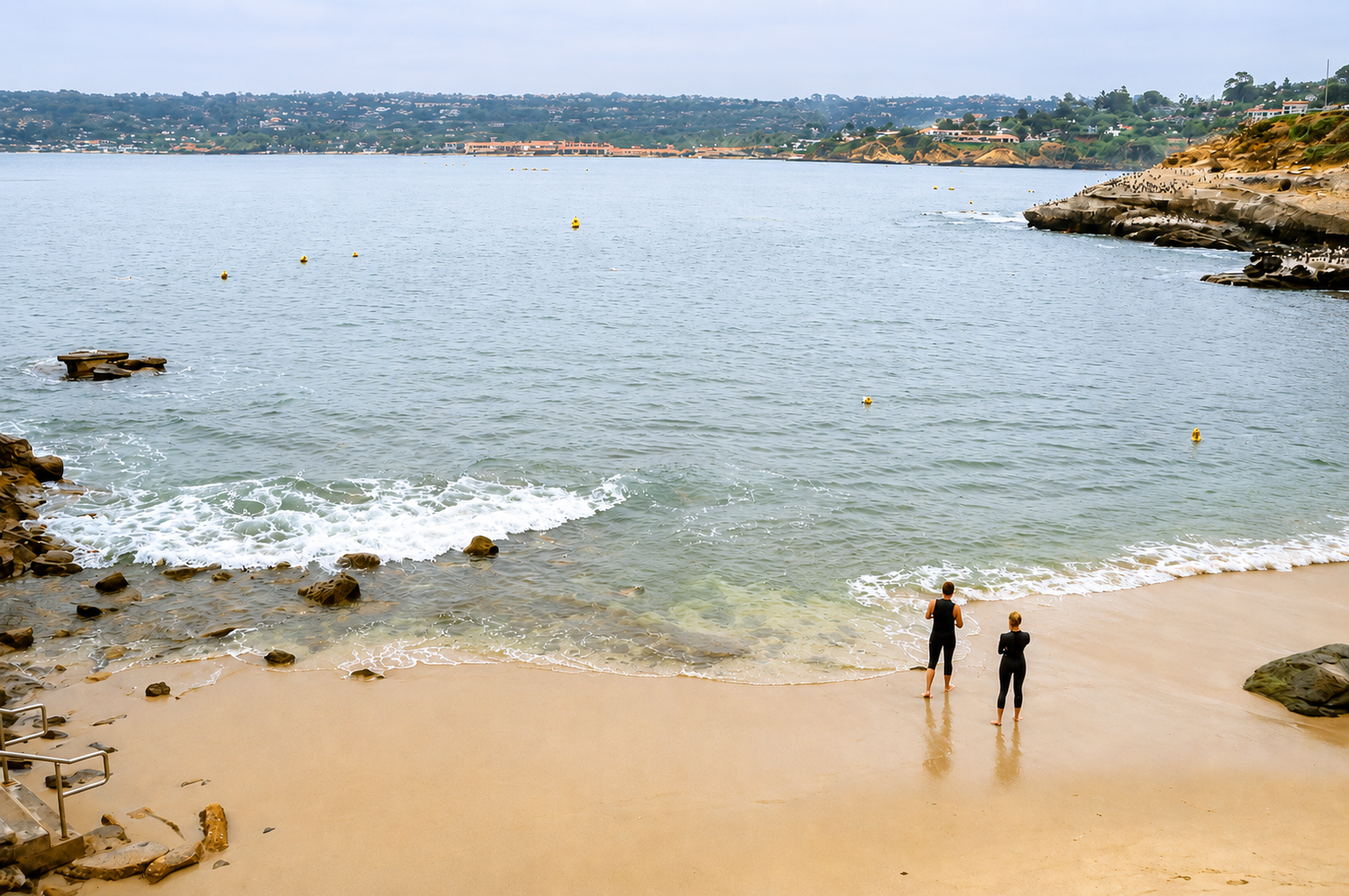 Two people in wetsuits stand on a sandy beach near the water while gentle waves roll in. Rocky outcrops frame the shore; a distant town on green hills lines the horizon under a cloudy sky.