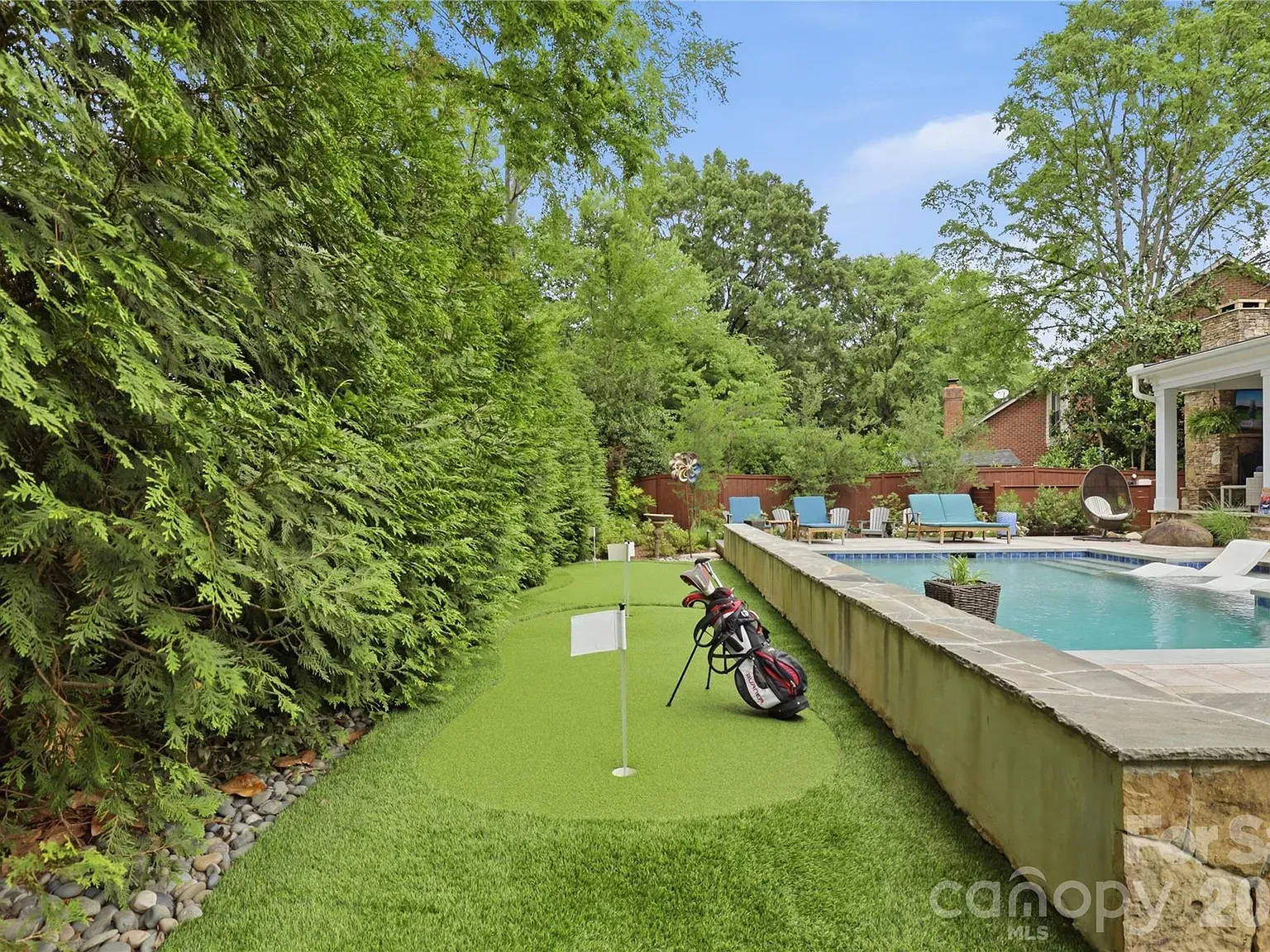 Backyard pool area with a small putting green, flag, and a golf bag with clubs on the grass. Blue lounge chairs surround the pool; tall hedges and trees frame the scene.