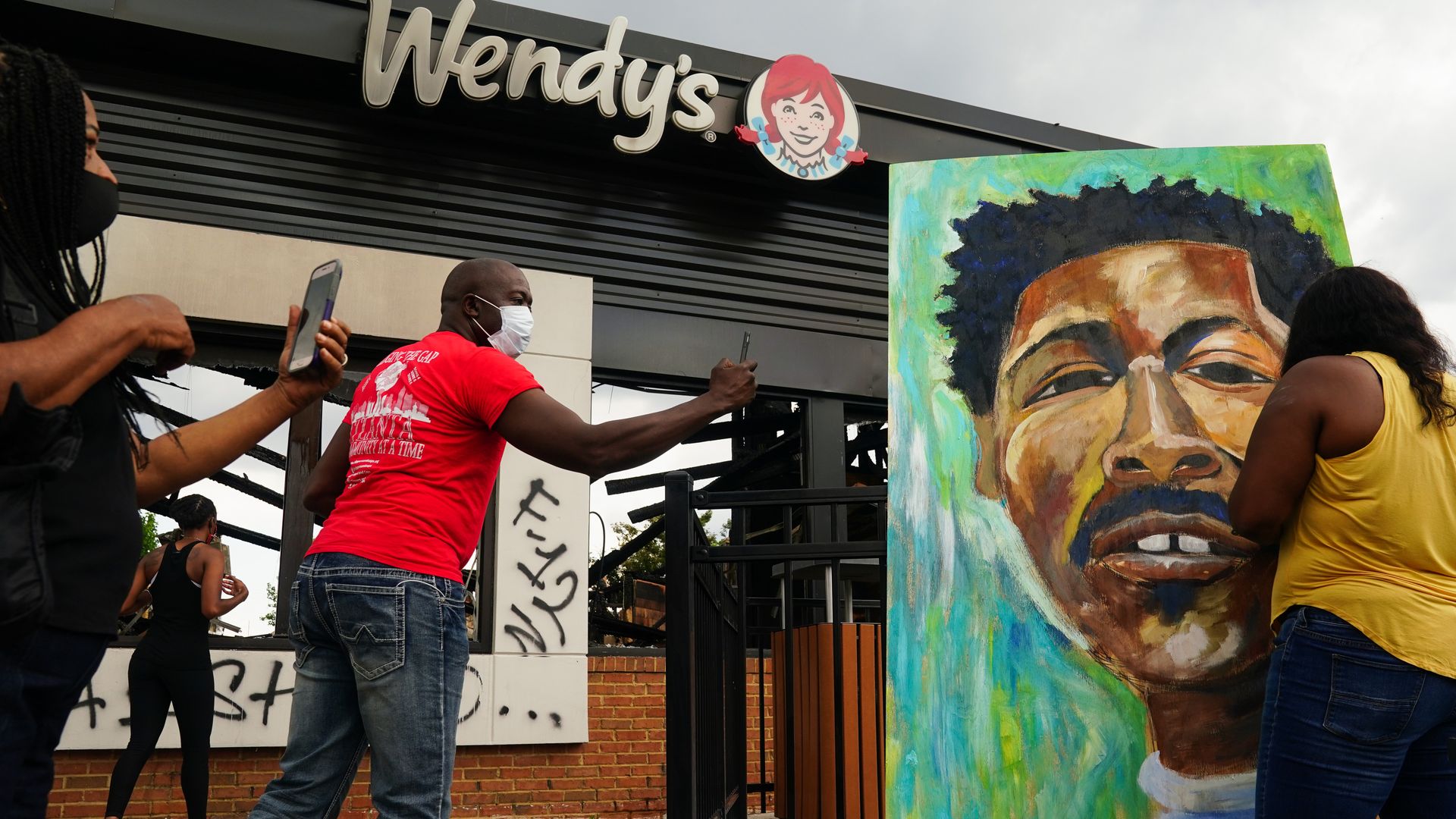 People use their cellphones to document a muralist painting the face of Rayshard Jones outside a burned Wendys restaurant