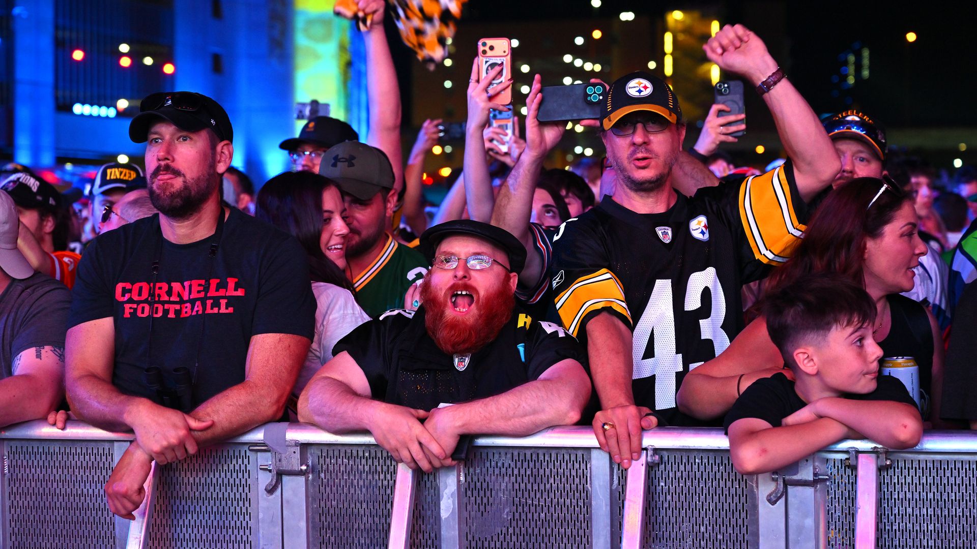 Nighttime stadium crowd behind a barrier, fans in black and yellow jerseys. Blue and colored lights illuminate the background as people hold up phones; one raises a tiger-striped scarf.