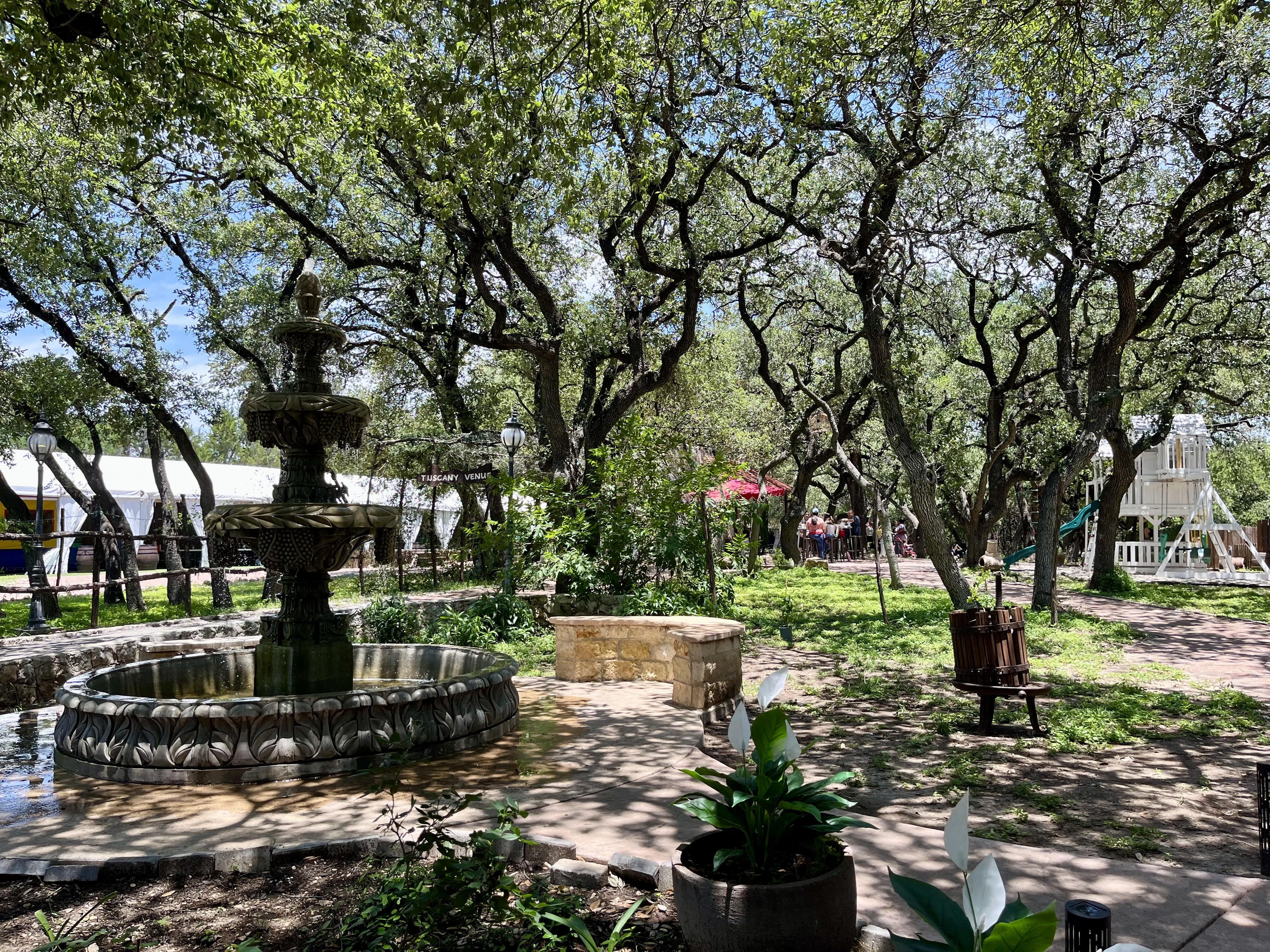 Sunlit courtyard with a large ornate fountain in the foreground, dense twisting trees forming a green canopy overhead; a sign reads Tuscany Venus near tents and people.