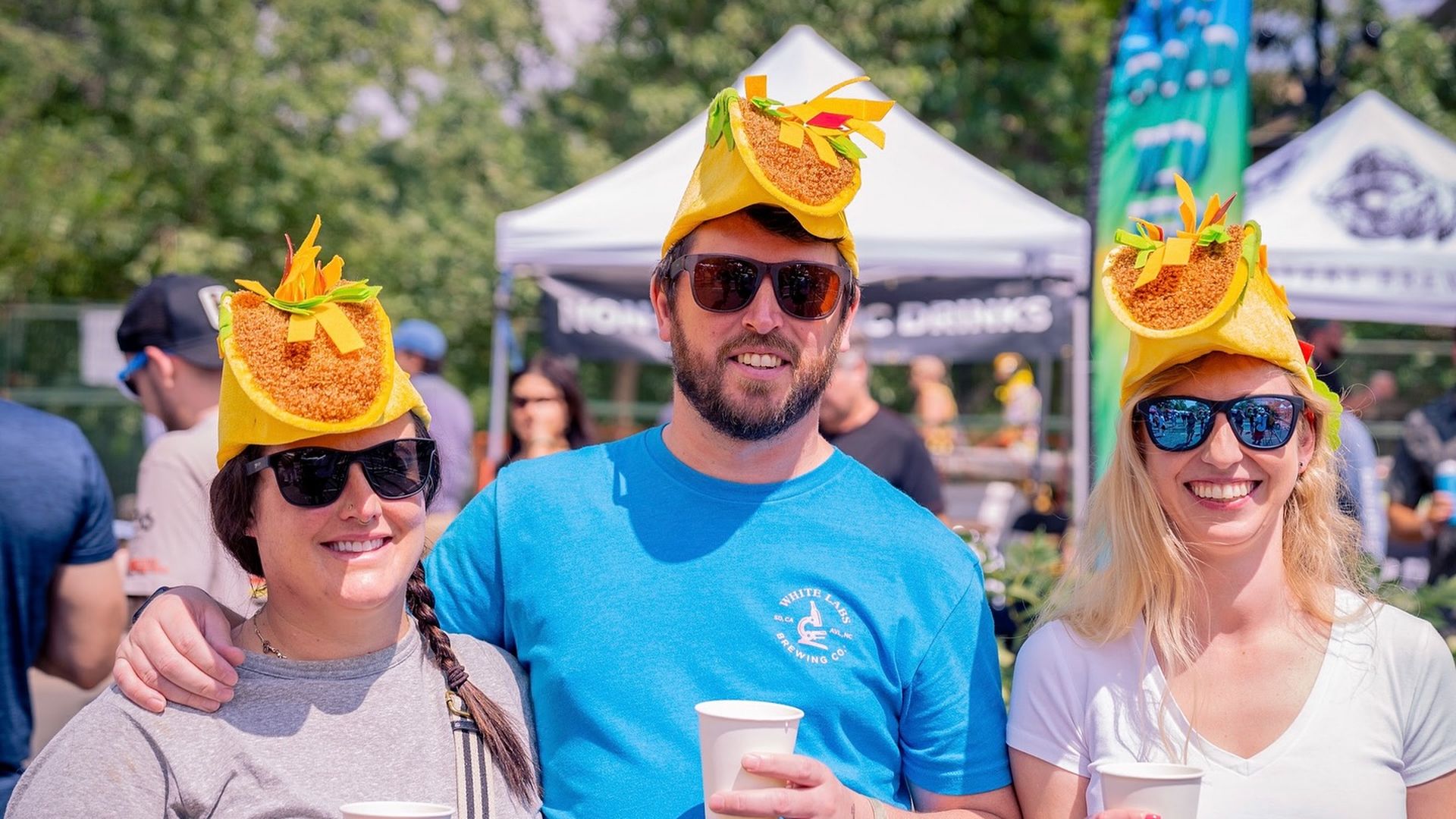 Three people wearing taco hats and sunglasses smile outdoors, each holding a white cup, with tents and trees in the sunny background.