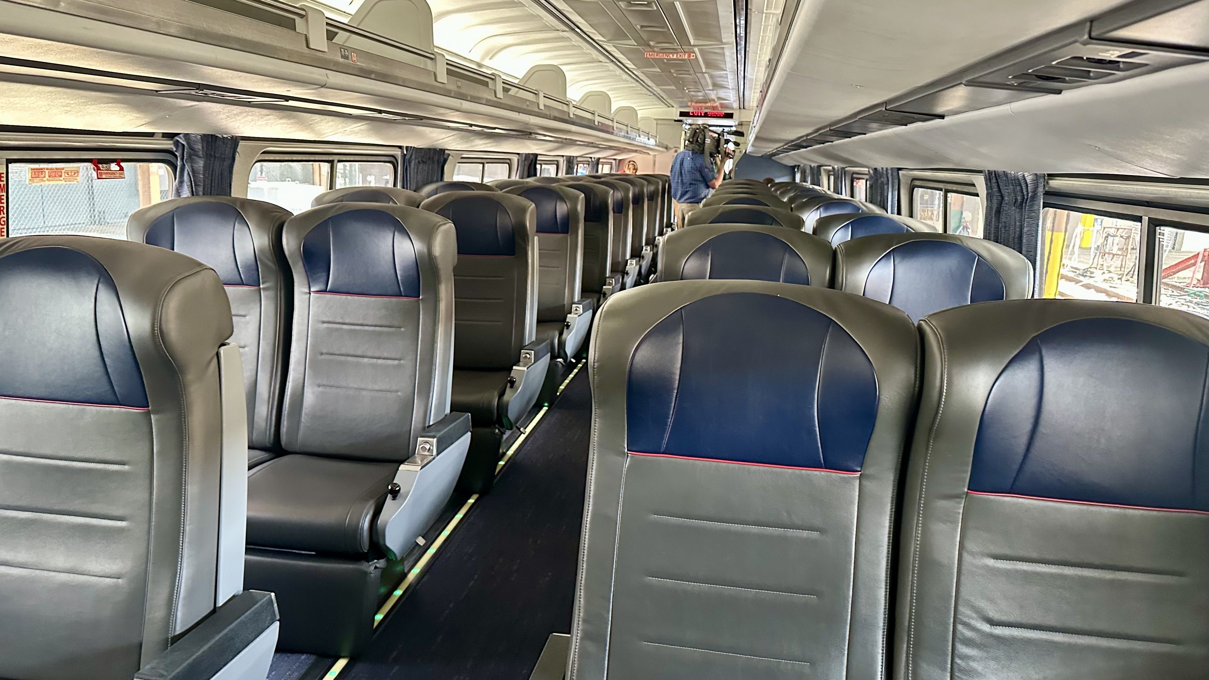 Interior of a clean, empty bus with rows of gray and blue leather seats, large windows with curtains, and a person standing near the rear holding a camera.