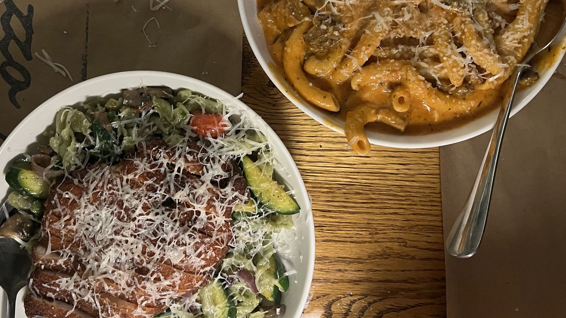 Two plates of pasta dishes on a wooden table. One plate has green pasta with grilled chicken, zucchini, mushrooms, and shredded cheese. The other has creamy orange pasta with mushrooms and cheese.