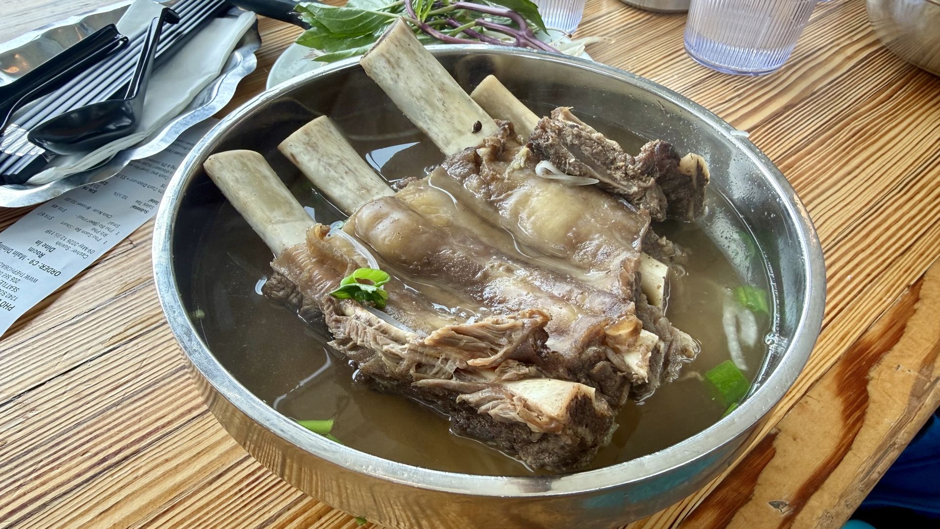 Metal pot with bone-in meat simmering in broth on a wooden table; a knife and utensils on a napkin, green leaves with purple stems, and ribbed clear plastic cups in the background.