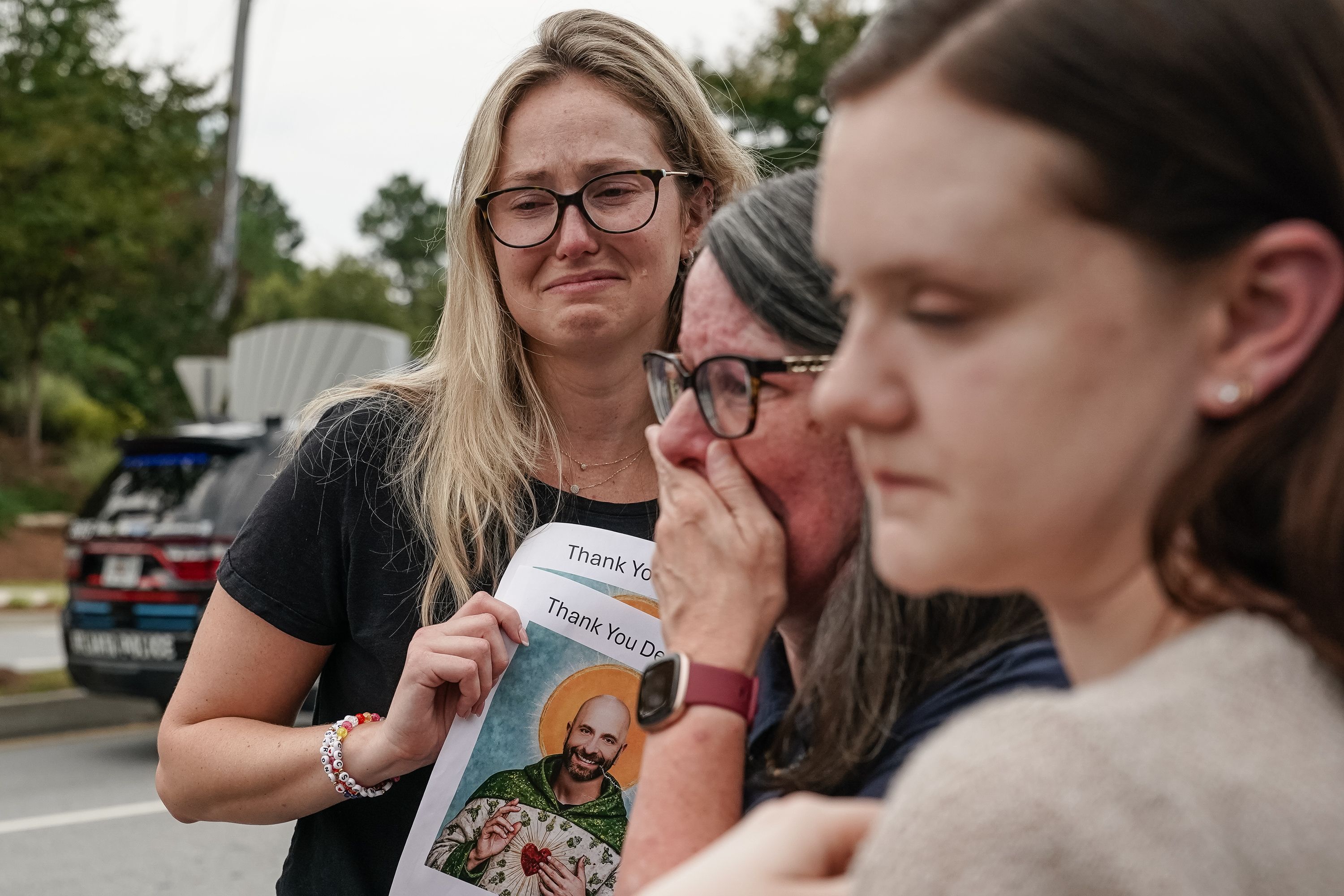 Three women outdoors, visibly emotional and crying; one holds a paper with a man's photo and text saying "Thank You". A police vehicle is visible in the background.