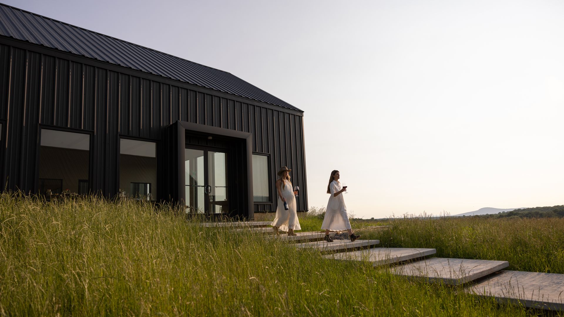Two women in white dresses walk down outdoor steps beside a modern black building, holding wine glasses and bottle, with tall green grass and a distant hill under a pale sky.