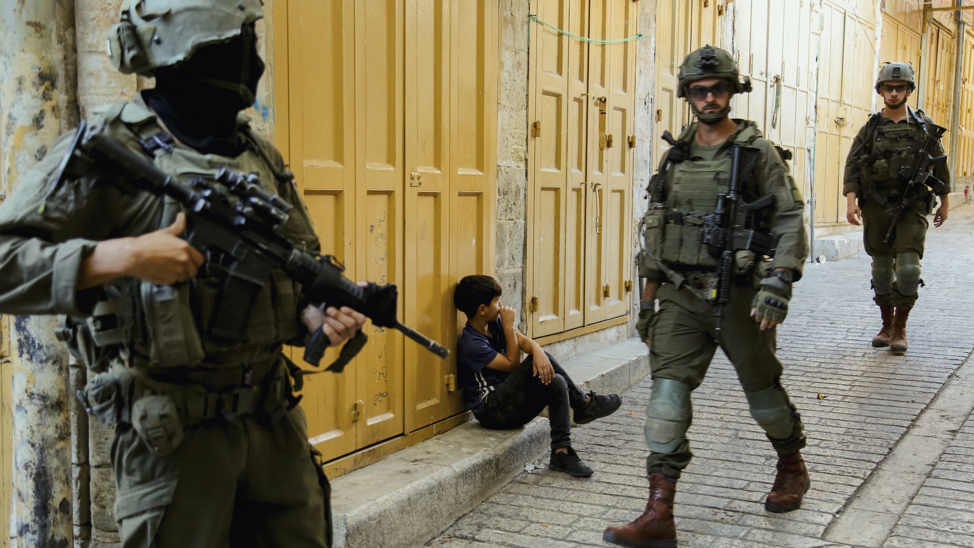 A boy sits in front of a closed shop in the Old City of Hebron, West Bank, on Oct. 18, 2025, as Israeli forces look on. 
