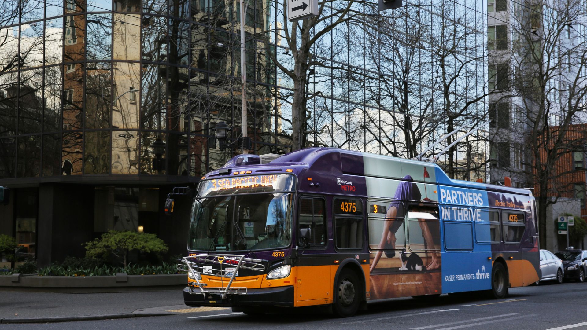 A purple-and-yellow King County Metro bus travels along a downtown Seattle street, with office buildings and leafless trees in the background.