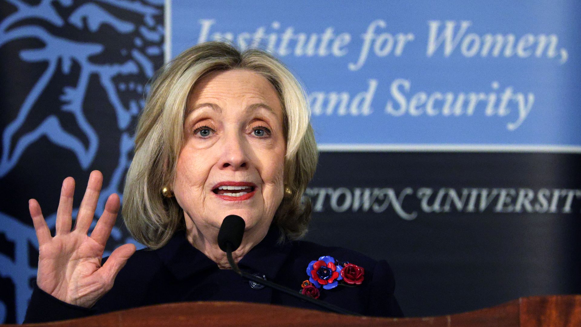 Woman with blonde hair and blue eyes speaking at a podium, wearing a dark outfit with red and blue flower pins, raising her left hand. Blue and black background with blurred text.
