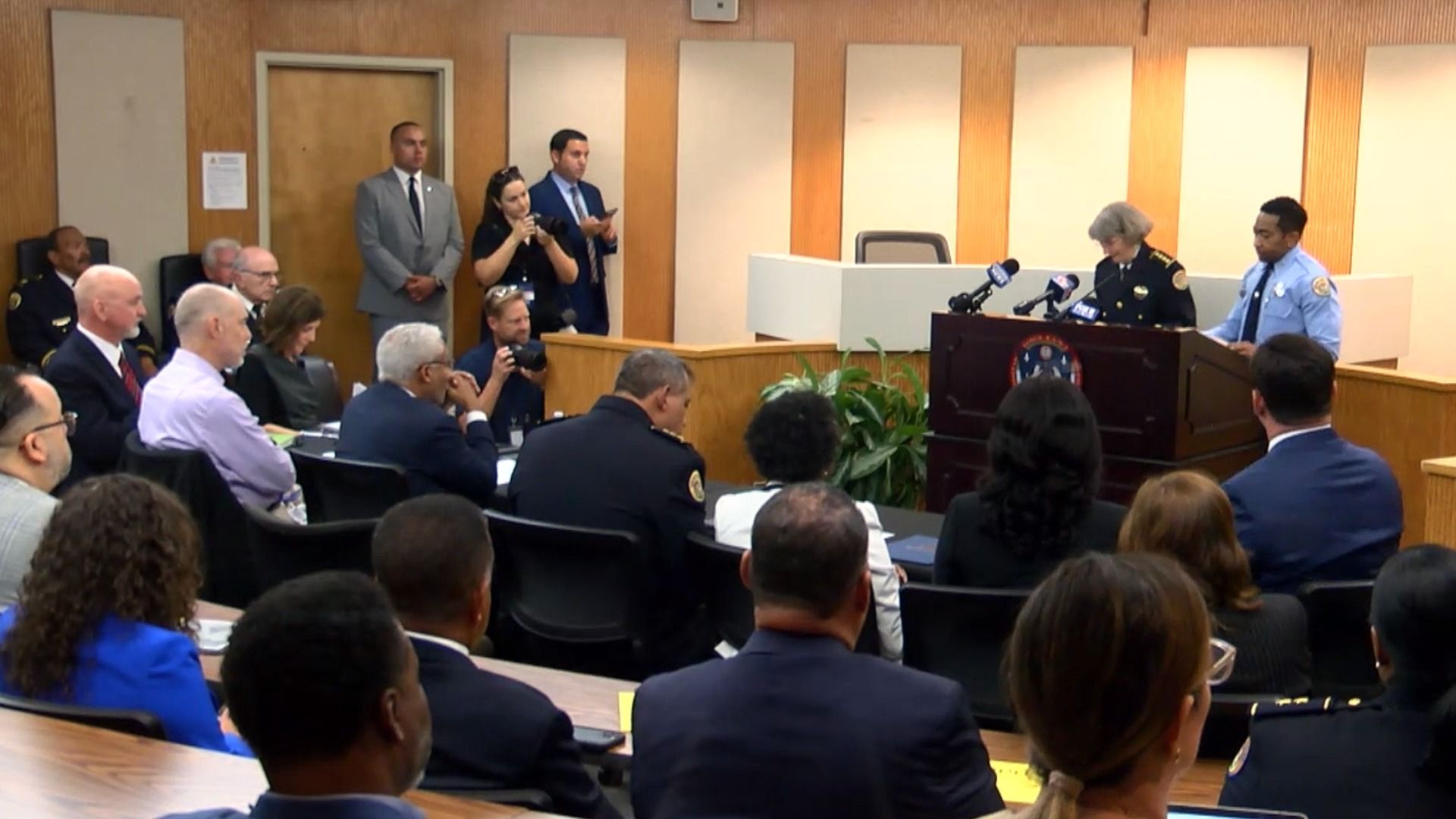 A police press conference in a wood-paneled room with officers speaking at a podium and an audience of officials and media listening and photographing the event.