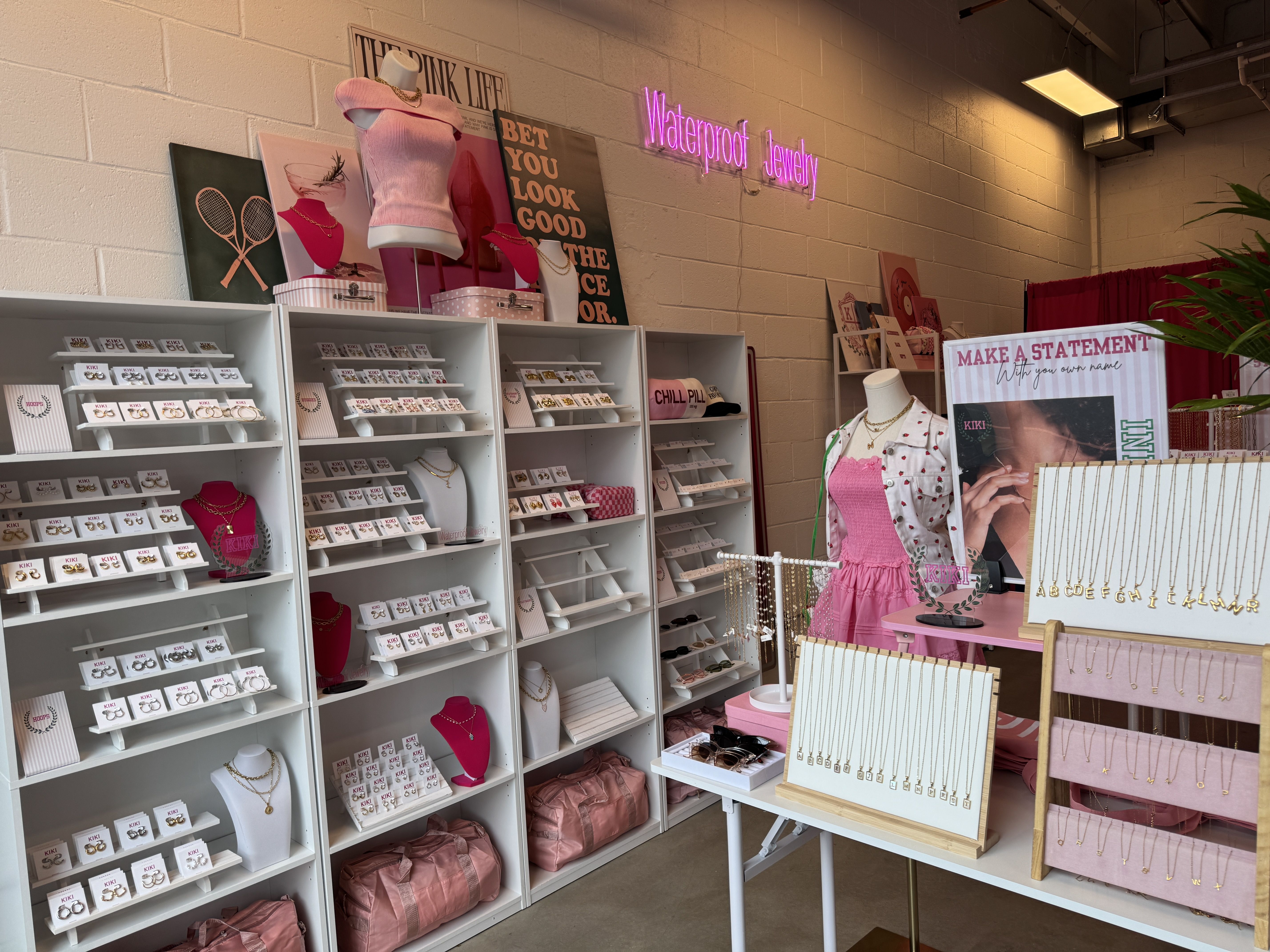 Jewelry shop display with white shelves and pink accents showing earrings, necklaces, and pink bags. Pink dress on mannequin, signs include "Waterproof Jewelry" in neon and "MAKE A STATEMENT".