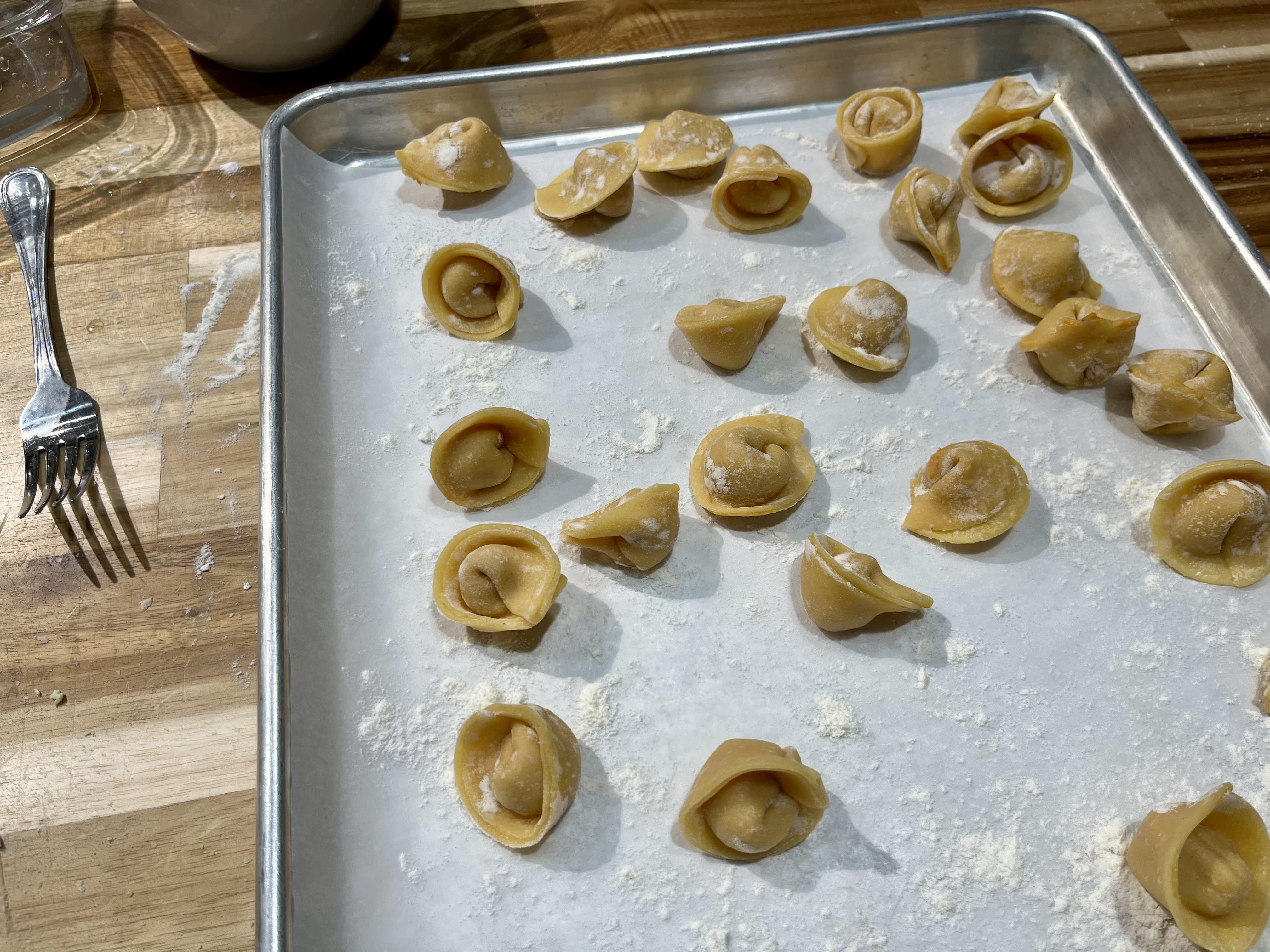 Uncooked, homemade tortellini on a floured metal baking sheet, arranged in scattered rows, on a wooden table. A fork sits to the left at the Central Market Cooking School in San Antonio.