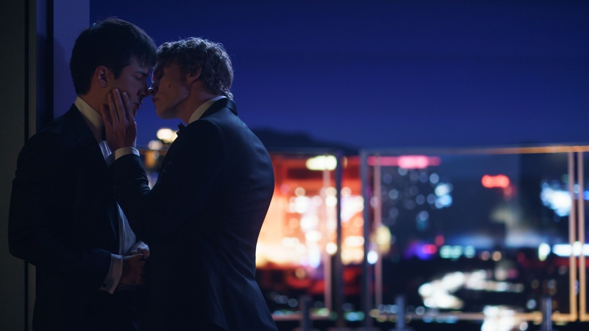 Two men in suits kiss against a wall on a rooftop at night in a city. 