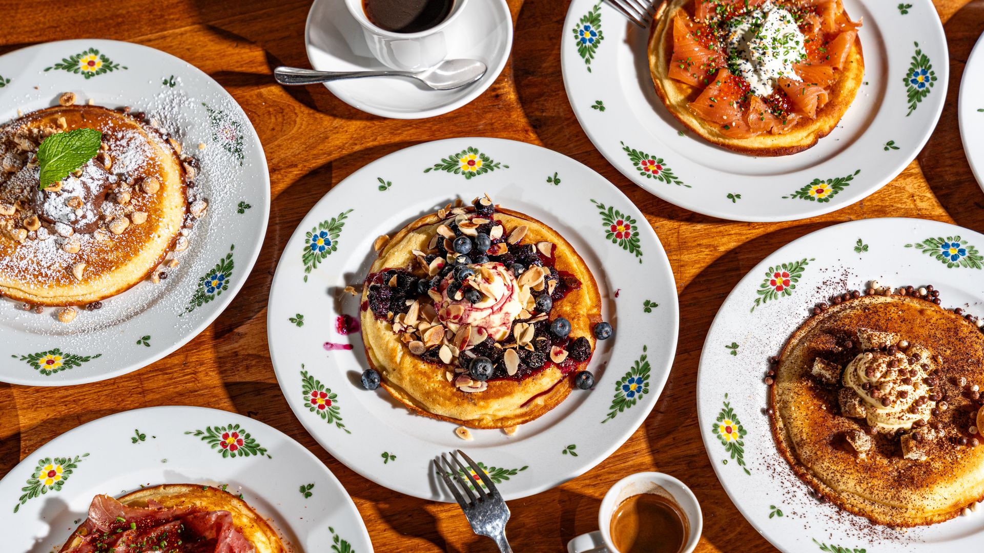 Five plates of assorted pancakes with toppings including blueberries, nuts, cream, smoked salmon, and ham, arranged on a wooden table with two cups of coffee.