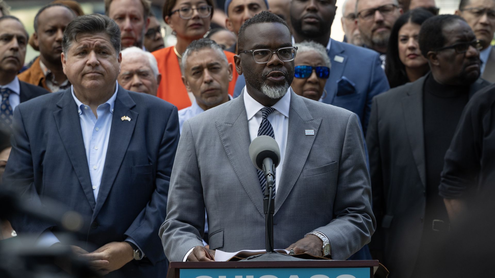 Man in gray suit with glasses speaking at a podium labeled "Chicago," surrounded by a diverse group of serious-faced people outdoors.