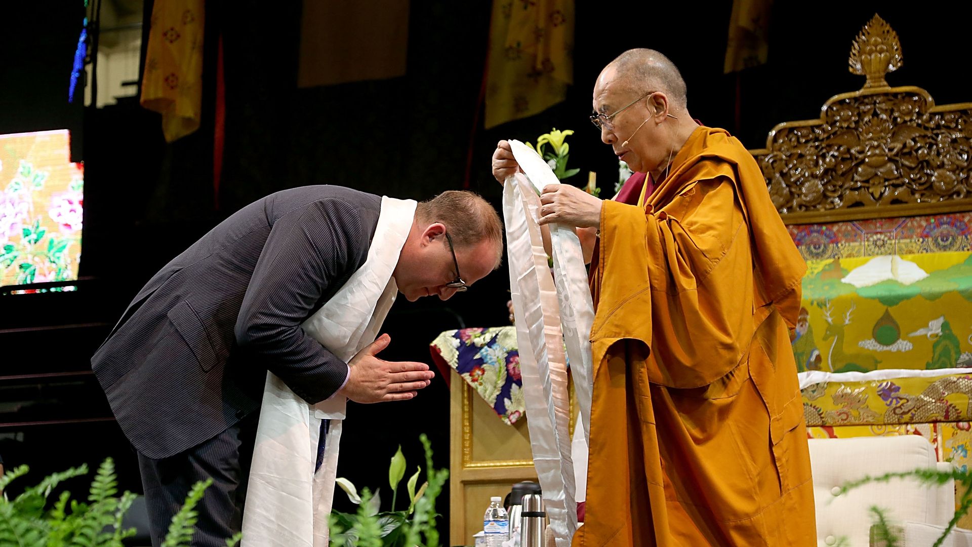 The Dalai Lama in orange robes presents a white ceremonial scarf to a bowed Jared Polis; ornate throne, bright backdrop, and greenery surround the scene.