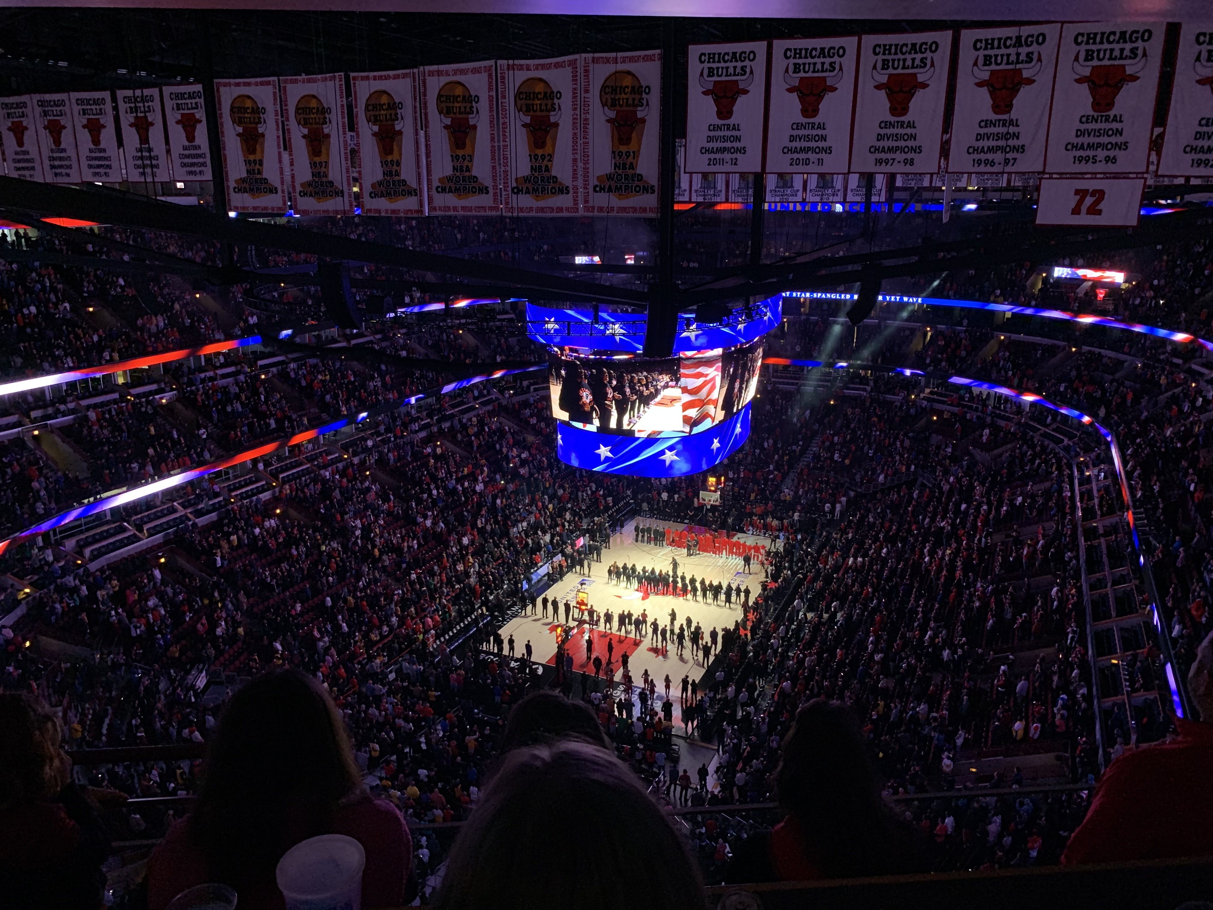 Overhead view of a packed Chicago Bulls basketball arena during player introductions, with teams lined up on the court and banners hanging above, featuring red, black, and blue lights.