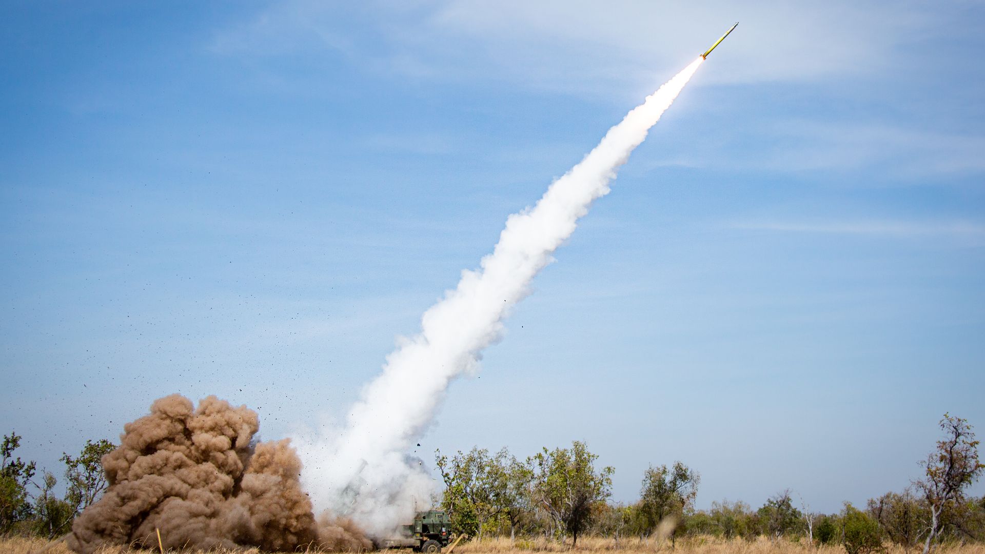 A rocket blasts off from a ground launcher, leaving a thick white smoke trail diagonally across a blue sky.